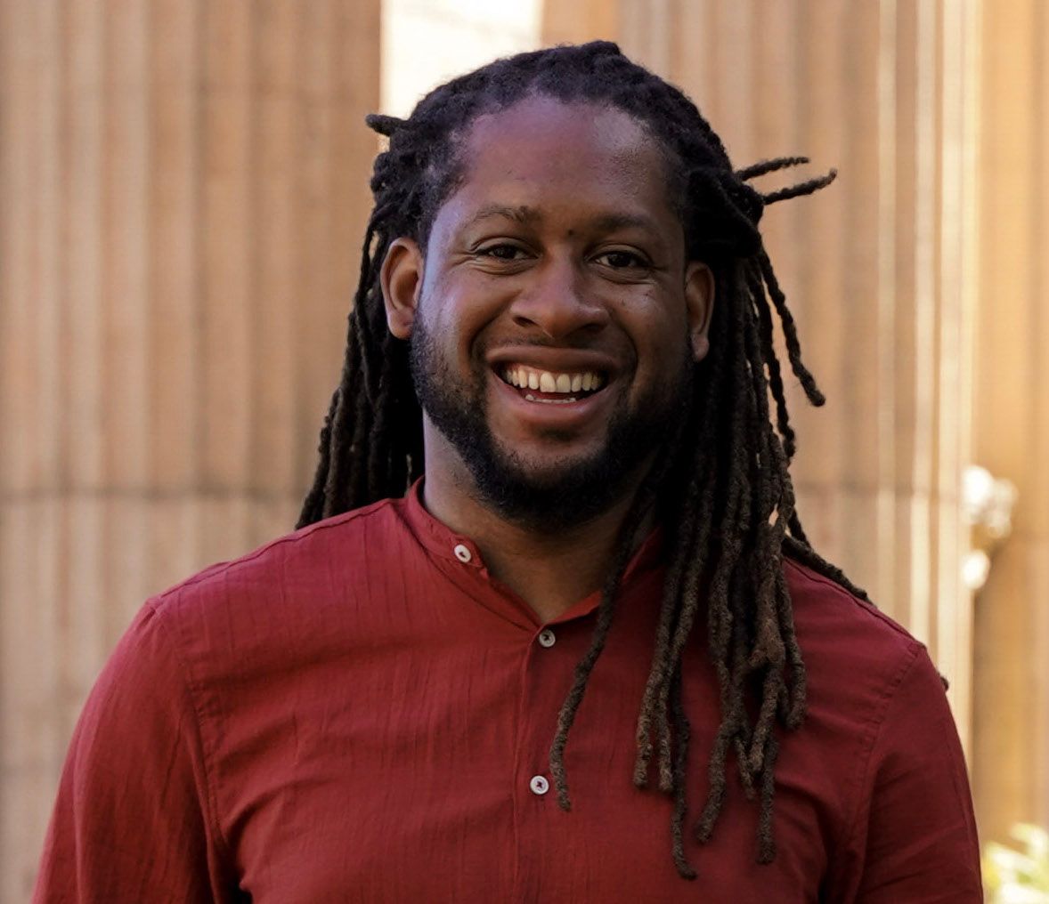 Smiling man with dreadlocks wearing a red shirt standing outdoors near a wooden wall.