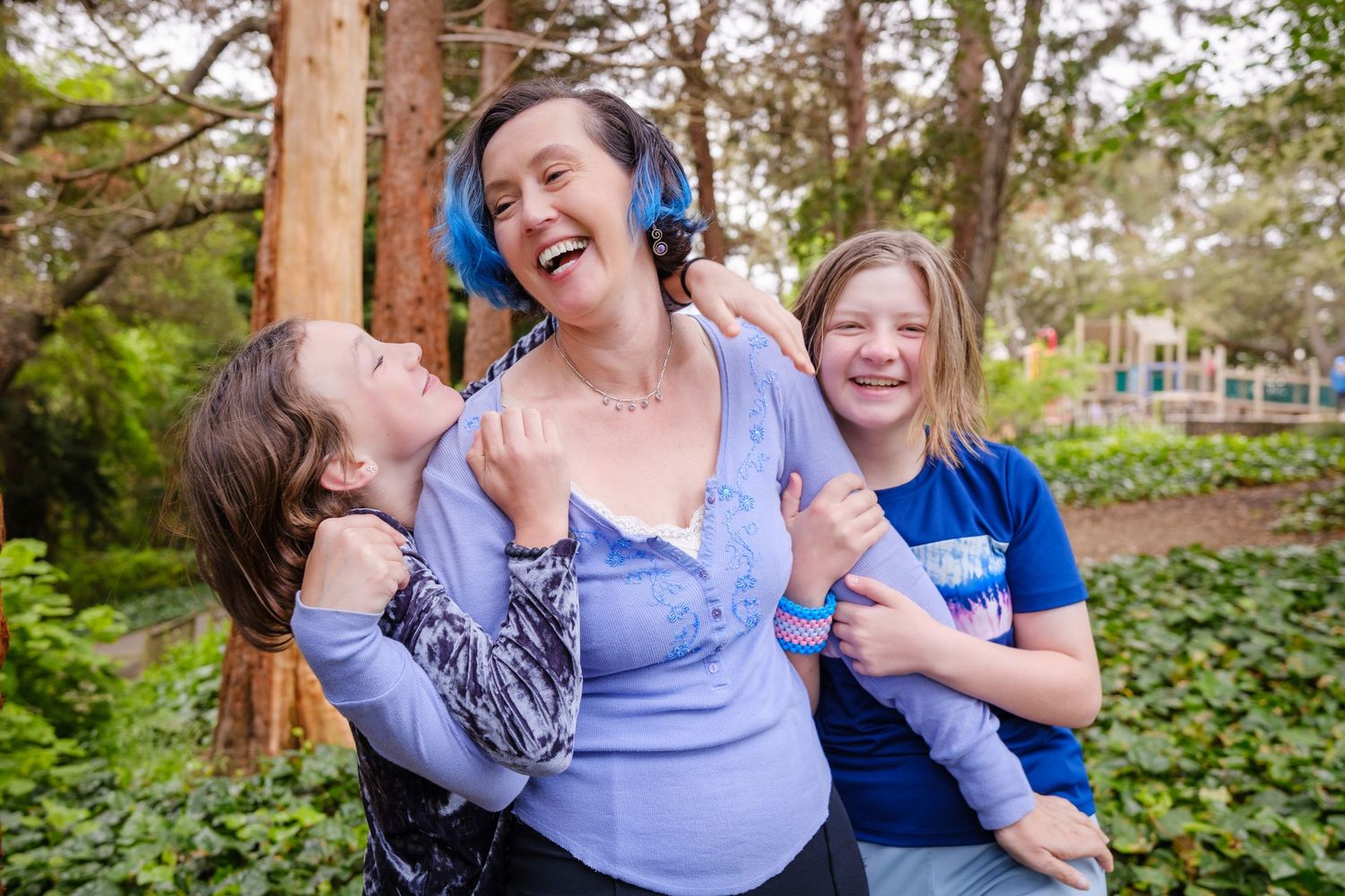 A laughing woman wearing blue headphones hugs two happy children outdoors in a lush green park setting.