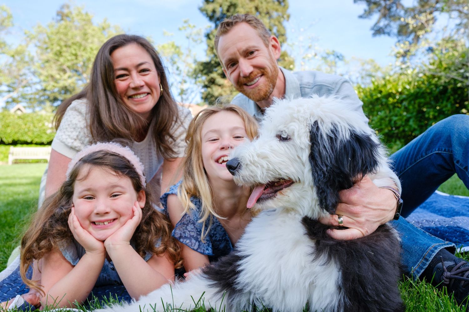 A happy family lying in the grass outdoors with their smiling Old English Sheepdog on a sunny day.