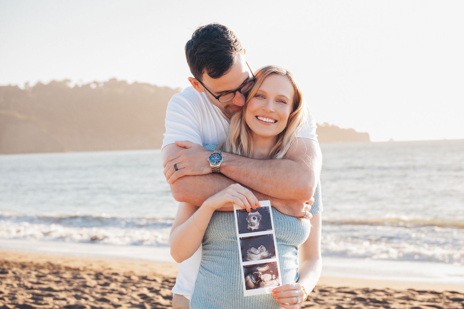 Couple on beach holding ultrasound photos, man embracing smiling woman, pregnancy announcement by the ocean.