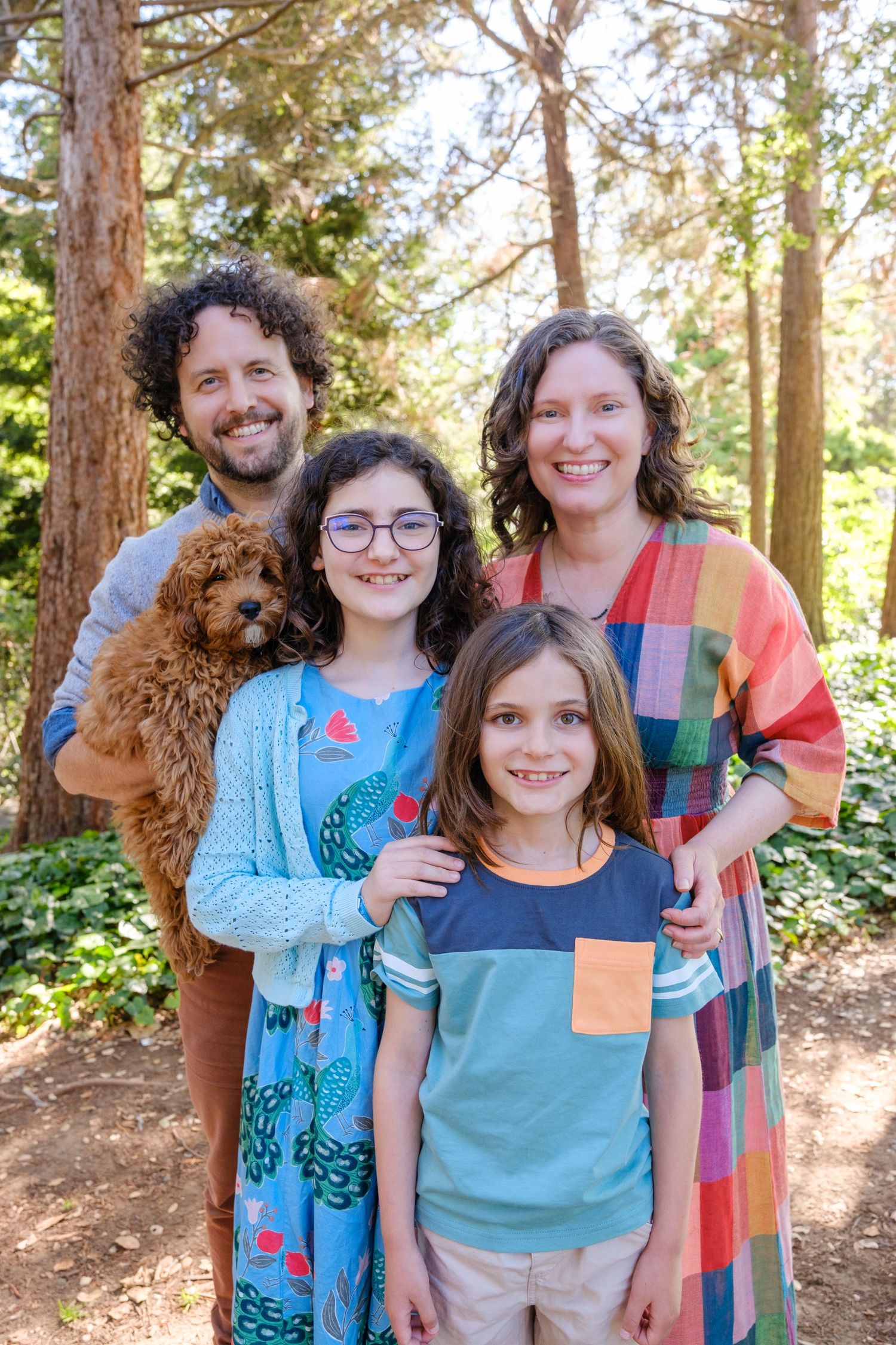 Happy family of four with a fluffy red doodle dog posing outdoors in a sunlit forest setting.