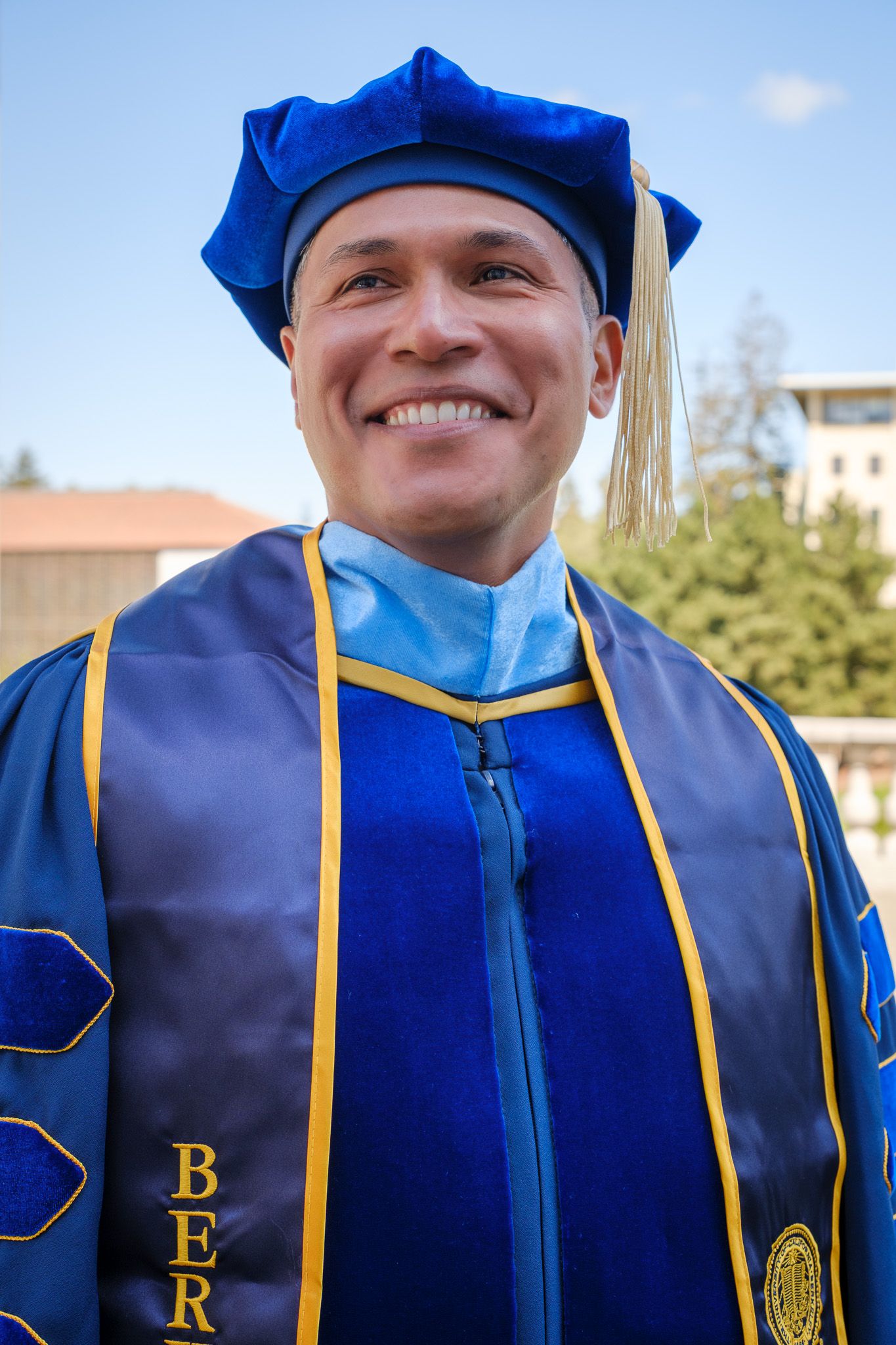 Smiling graduate in blue doctoral regalia and cap with gold tassel at UC Berkeley commencement ceremony.