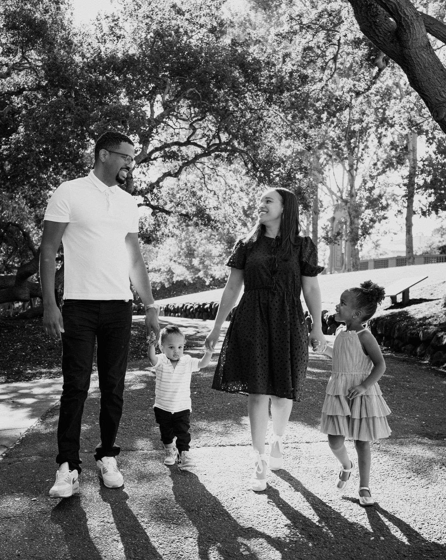 Family holding hands and walking together through a sunny park captured in black and white.