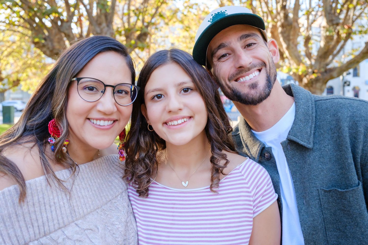 Three smiling people pose together outdoors, with trees and sunlight in the background.