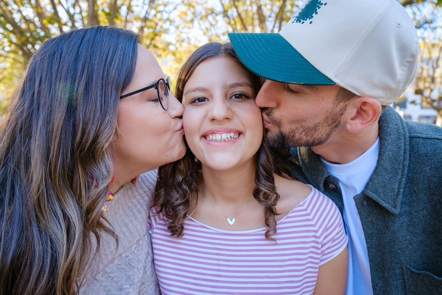 Parents kissing their smiling preteen daughter on both cheeks in a warm outdoor family portrait.