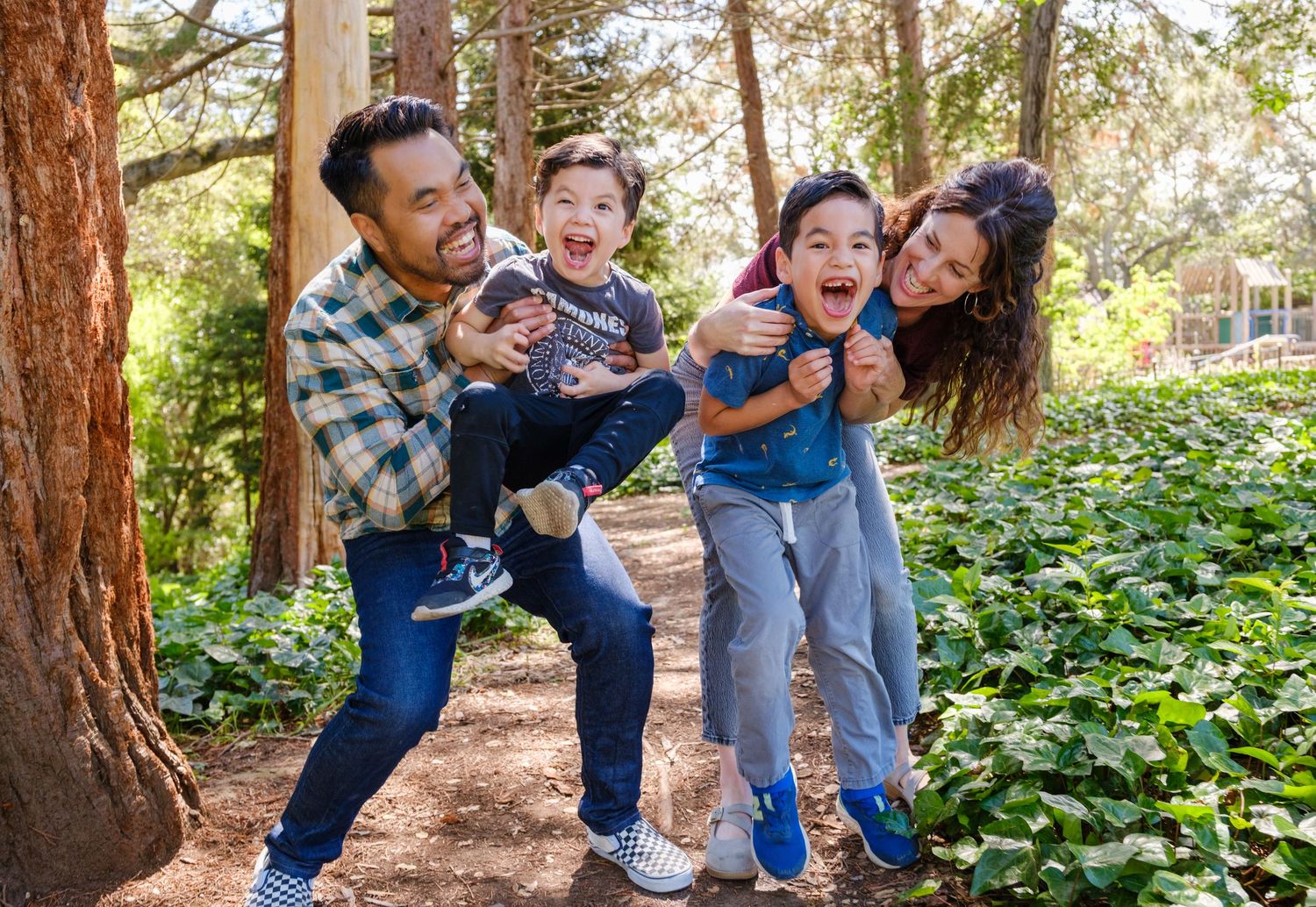 Family of four laughing and playing outdoors in a lush green park, parents carrying excited children.
