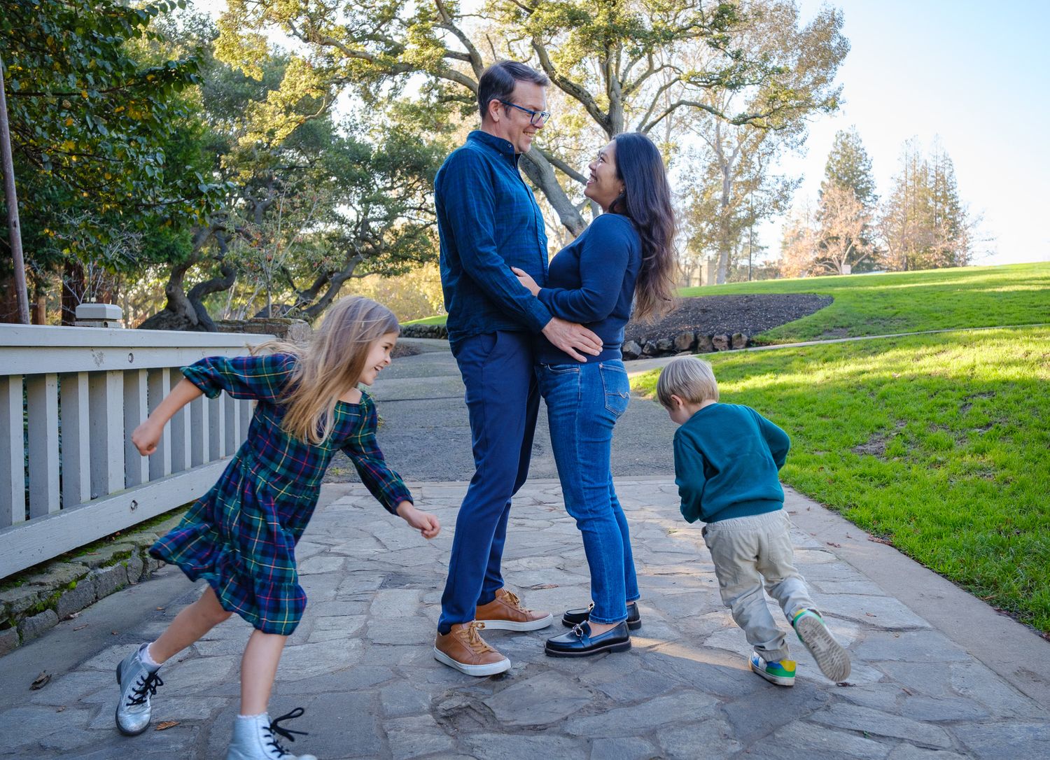 Family of four posing outdoors on stone path, parents embracing while two young children play around them in a park.