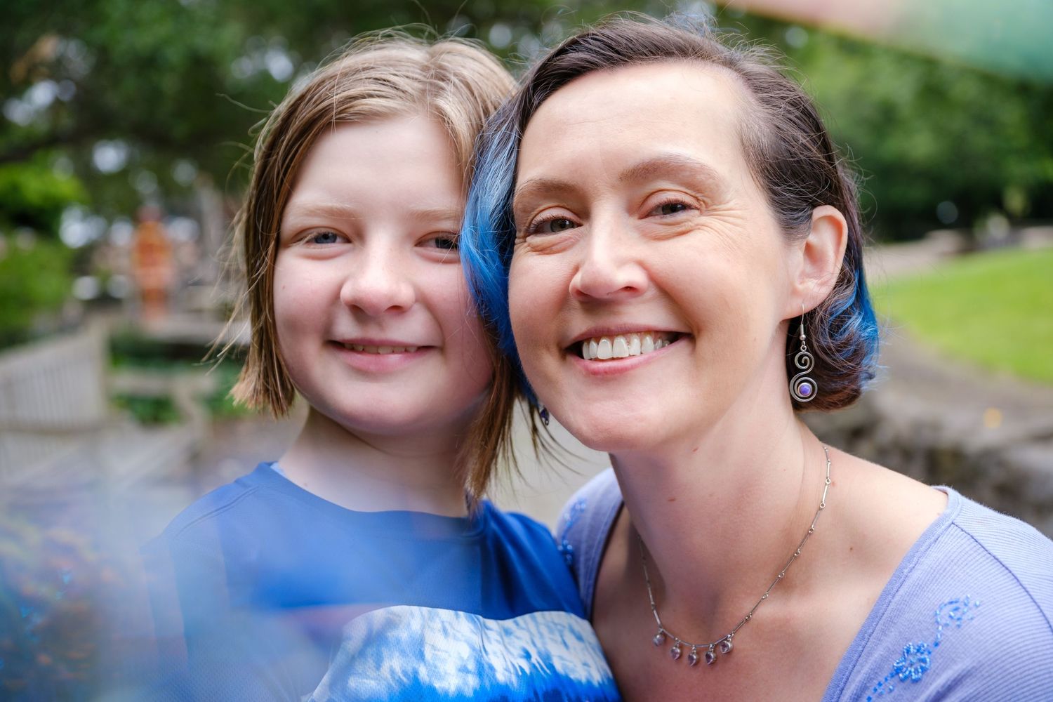 A smiling woman and young girl with blue-highlighted hair pose together outdoors in a lush green garden setting.