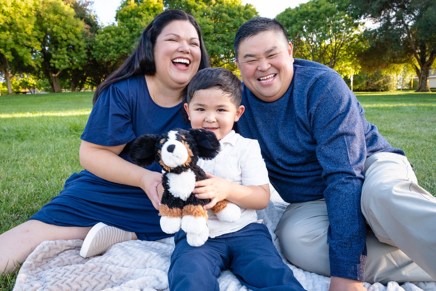 Happy family in matching navy blue outfits sits on picnic blanket with small black and white dog in sunny park.