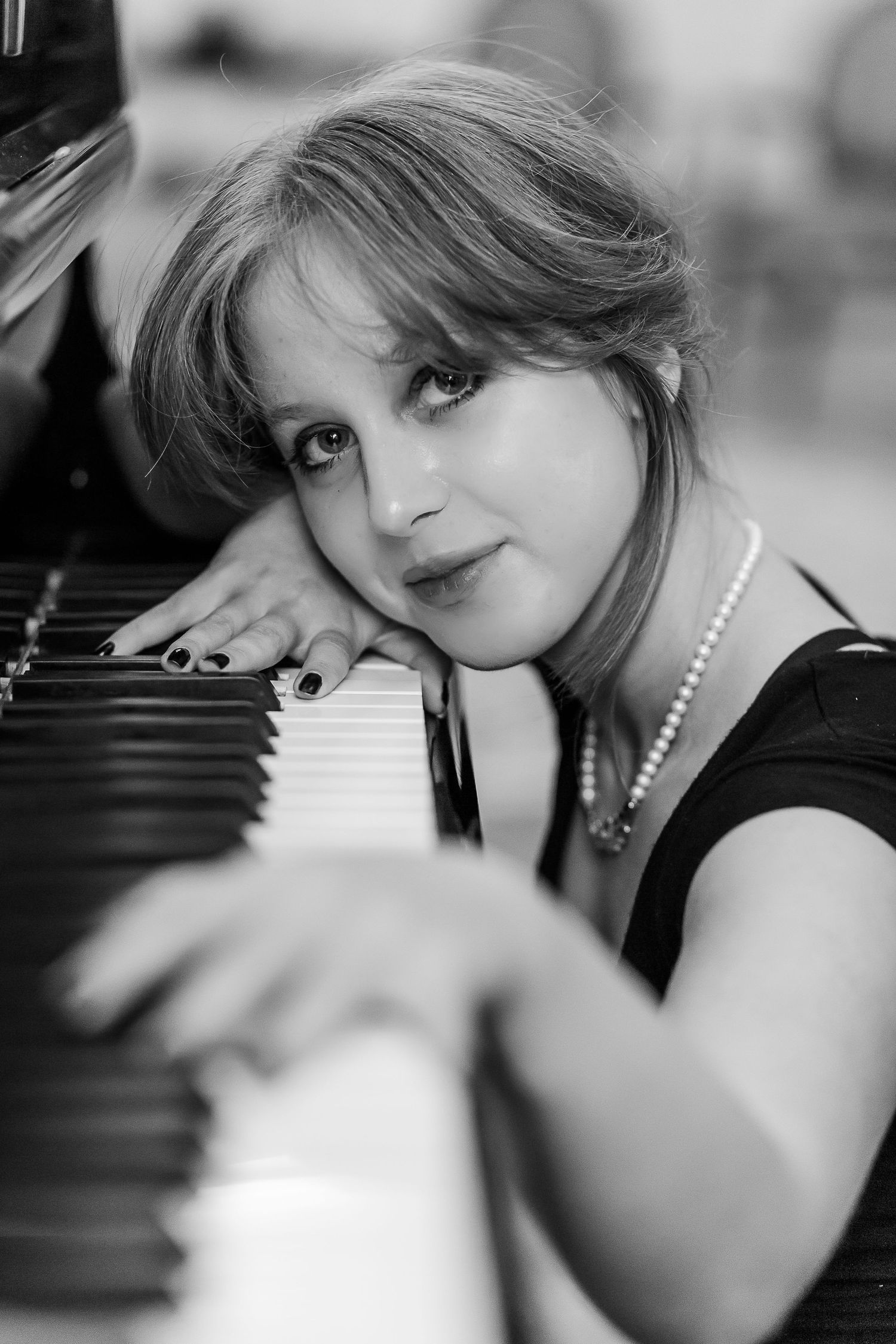Black and white portrait of pianist leaning against piano keys wearing pearl necklace and black sleeveless top.