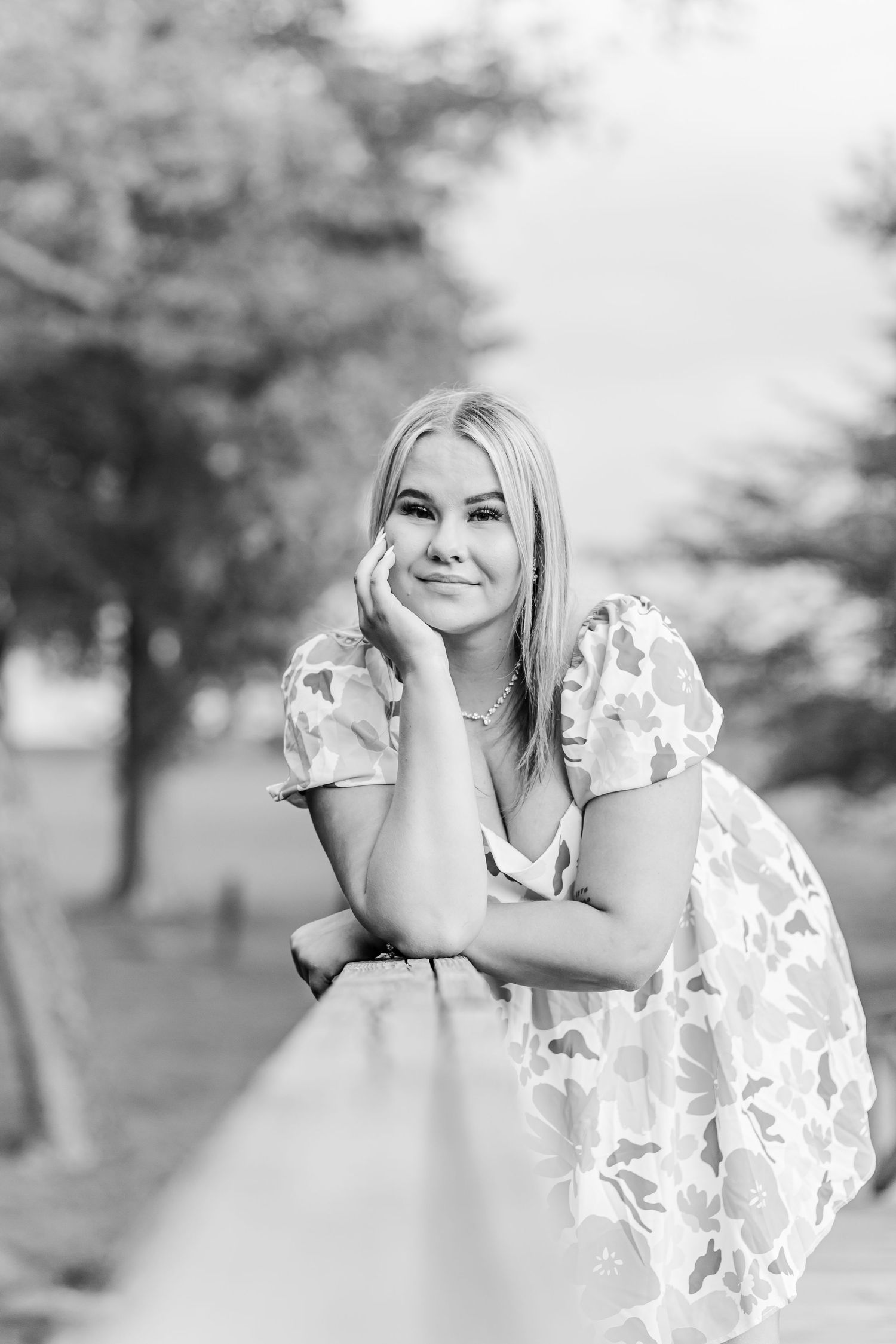 A black and white portrait of someone in a floral dress leaning on a railing outdoors with a joyful expression.