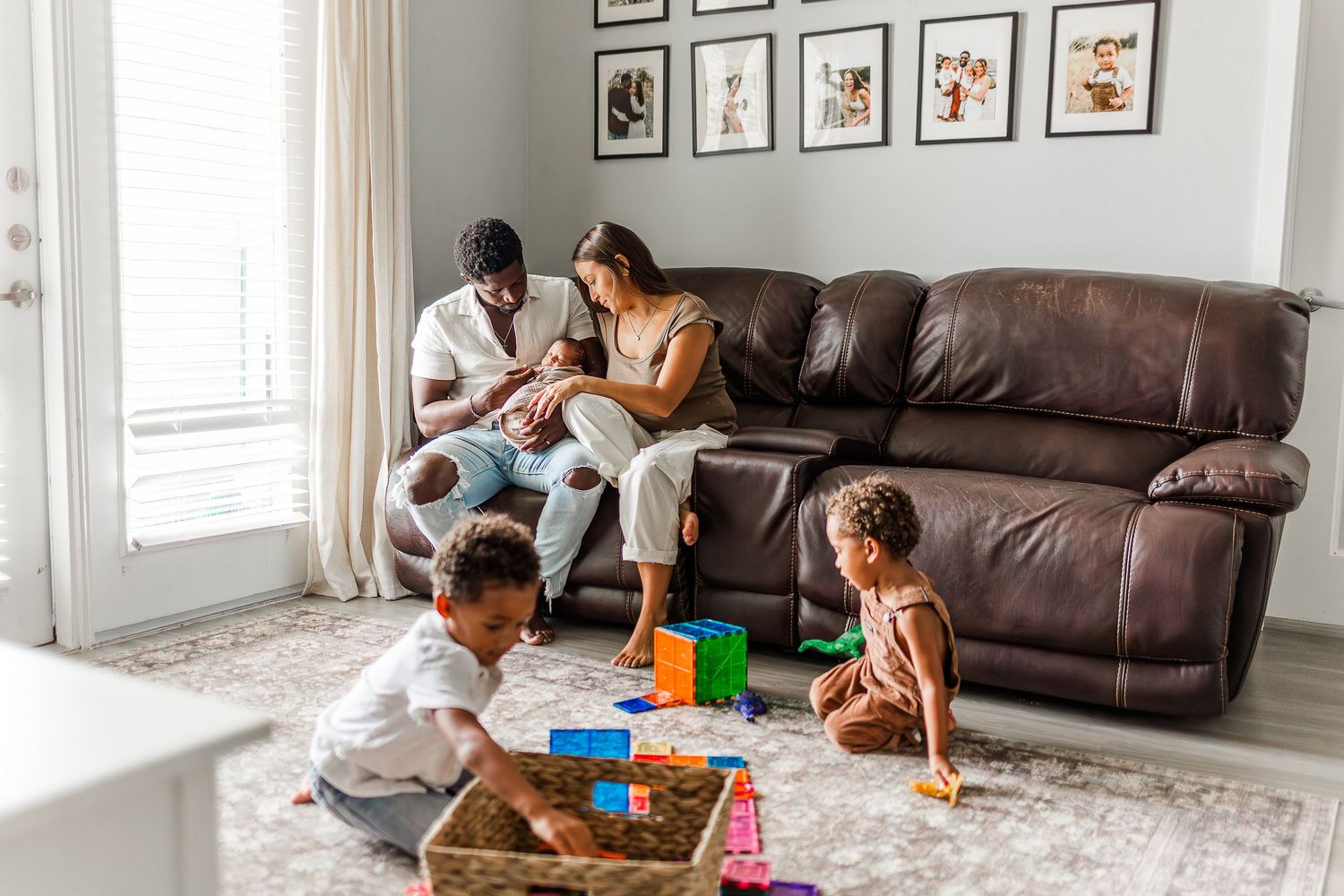 Family playing board games together on living room floor with brown leather couch and framed artwork on wall.