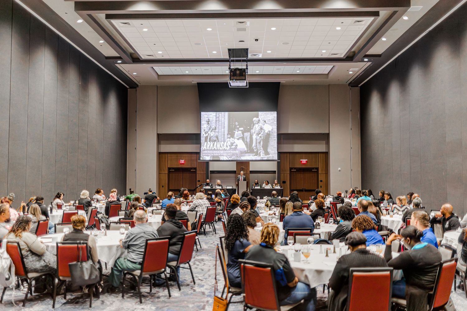 Conference attendees seated at round tables in a modern ballroom with a presentation screen displaying black and white footage.