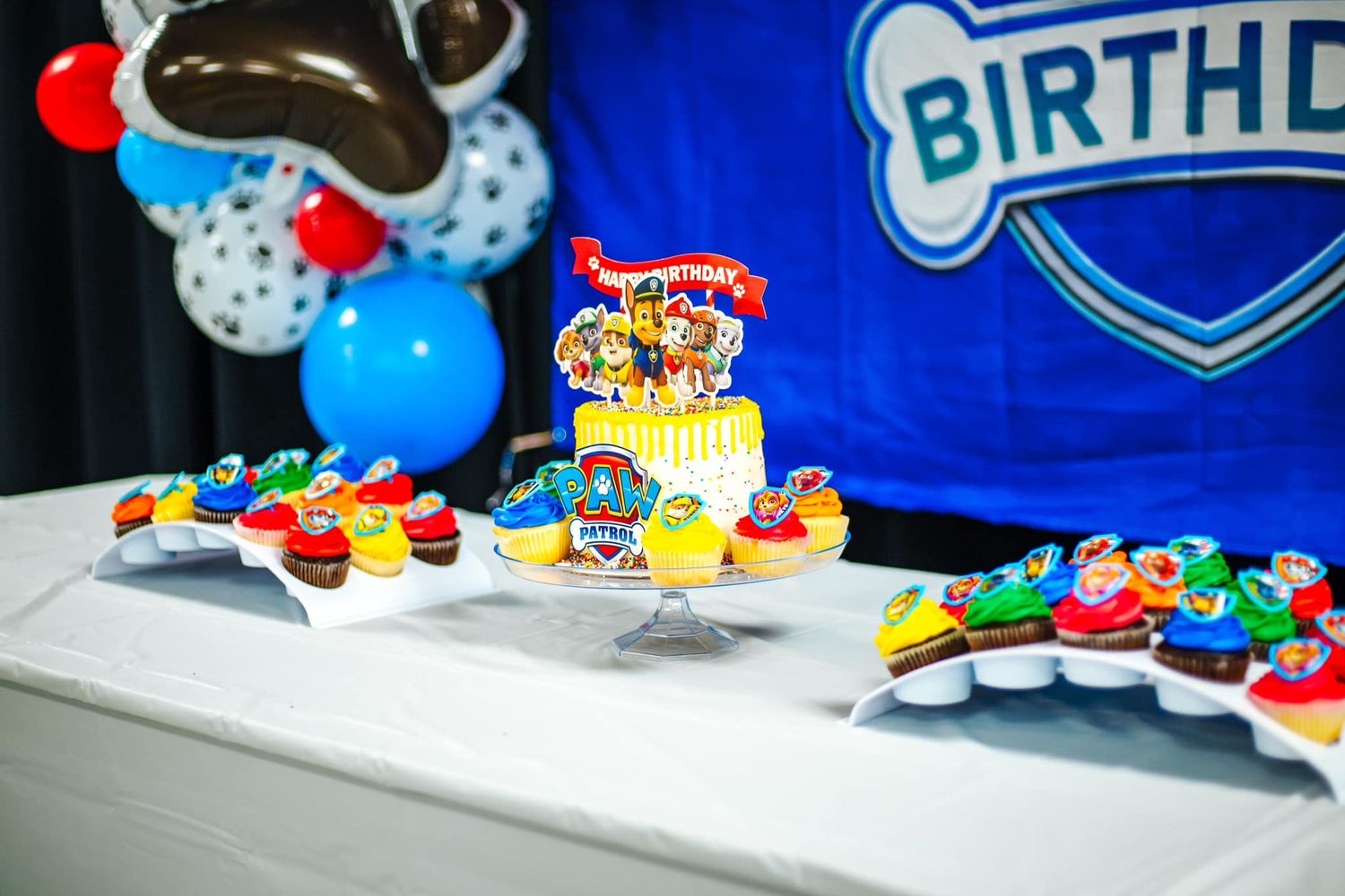 Colorful Paw Patrol themed birthday party setup with cake, cupcakes and blue balloon decorations on white tablecloth.