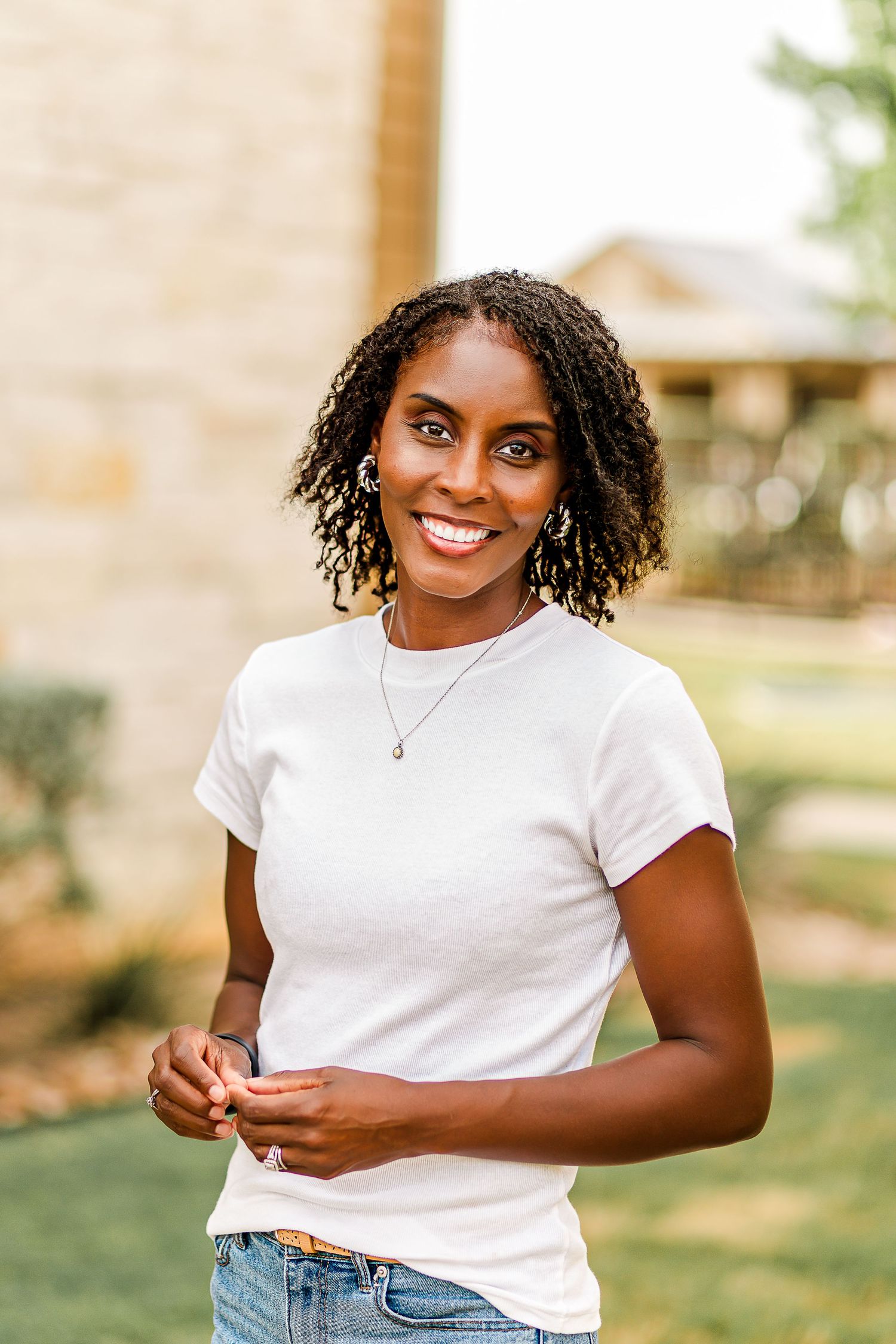 A person wearing a white t-shirt and jeans poses outdoors with natural lighting and a blurred background.