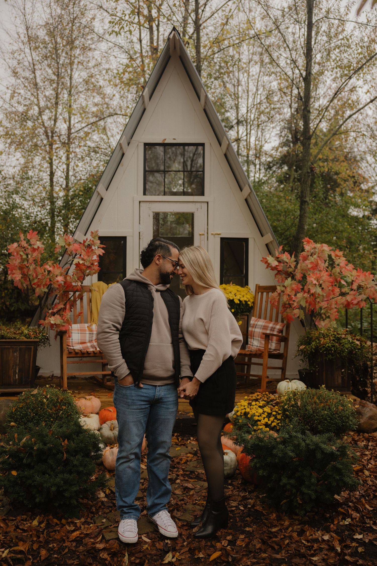 A-frame cottage with autumn decor featuring orange flowers, yellow mums and fallen leaves around the entrance.