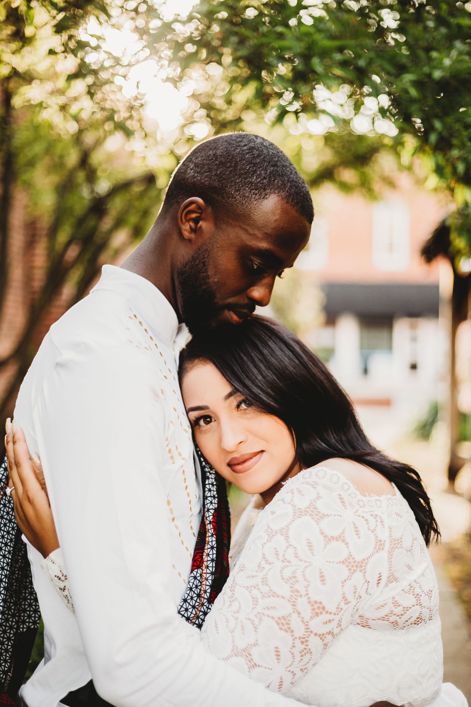 A romantic couple in white outfits embraces in a sunlit outdoor setting with trees and brick buildings in the background.