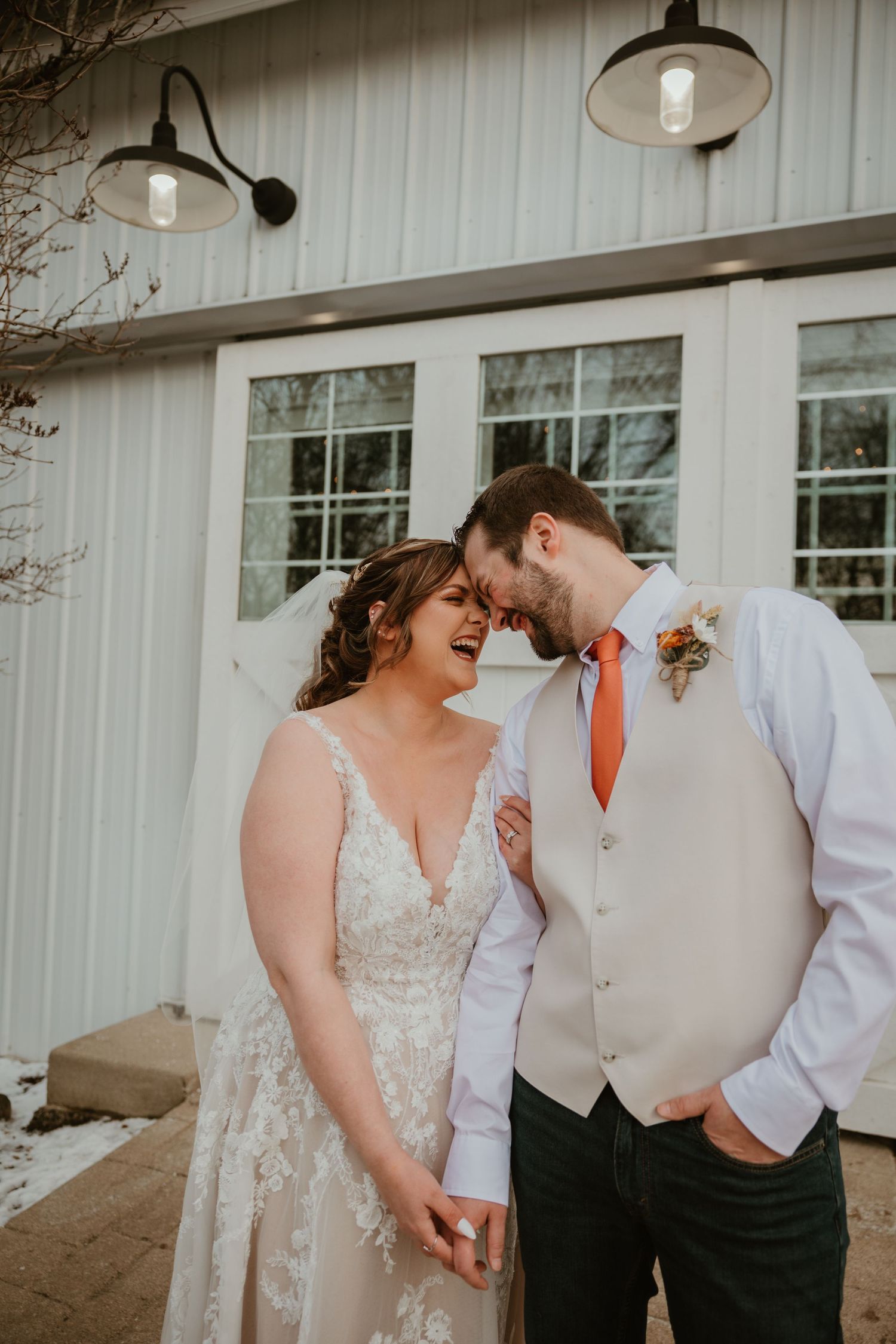 A joyful wedding couple share an intimate moment outside a white barn with vintage light fixtures.