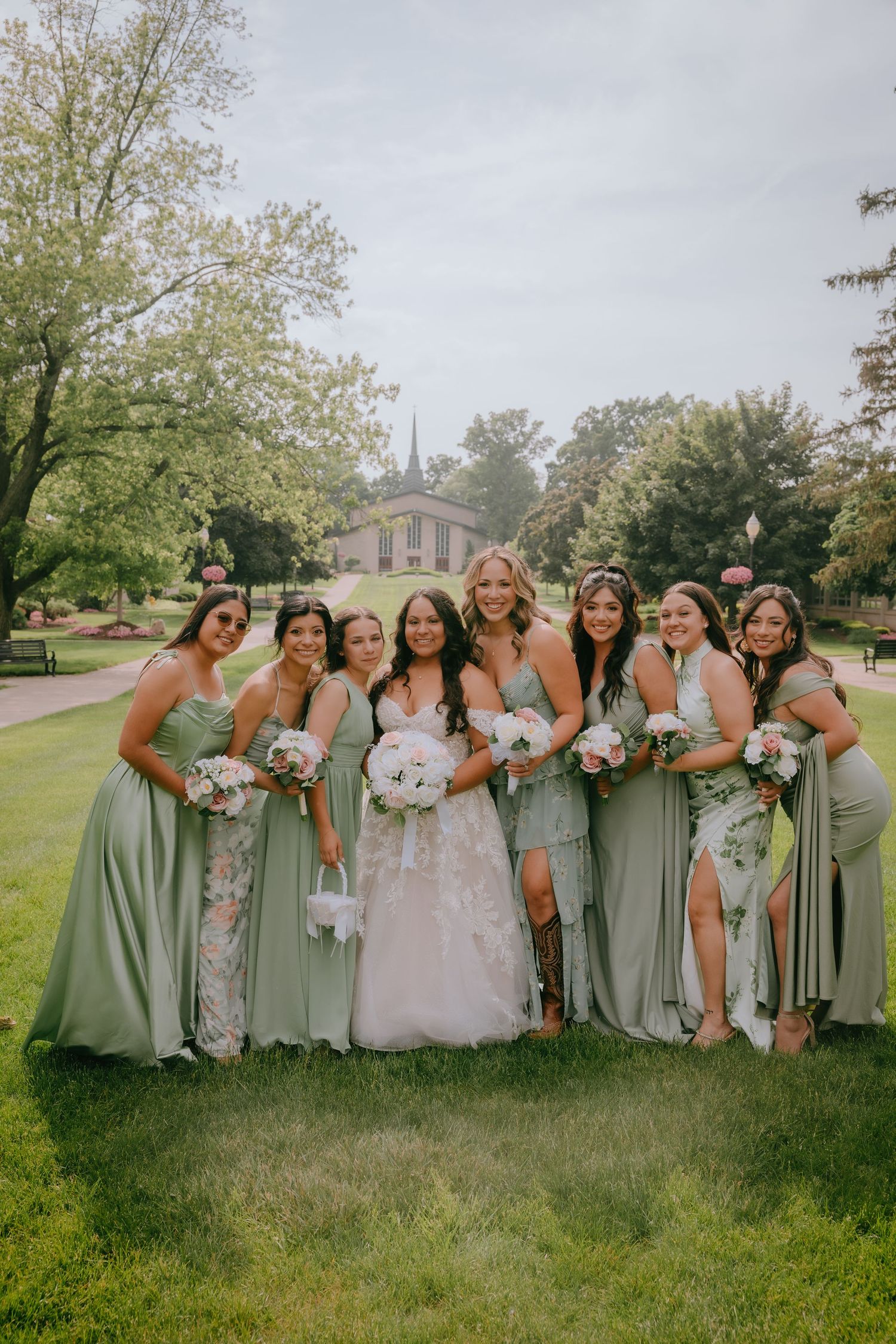 Wedding party wearing sage green bridesmaid dresses poses cheerfully on grassy lawn with bouquets.