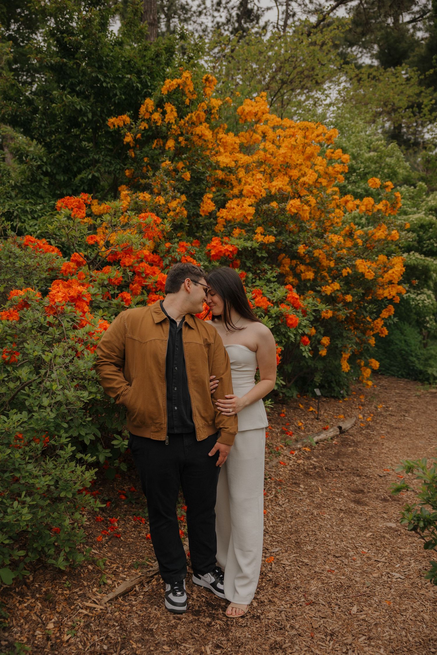 Couple sharing a romantic moment in a garden surrounded by vibrant orange and red flowering bushes.