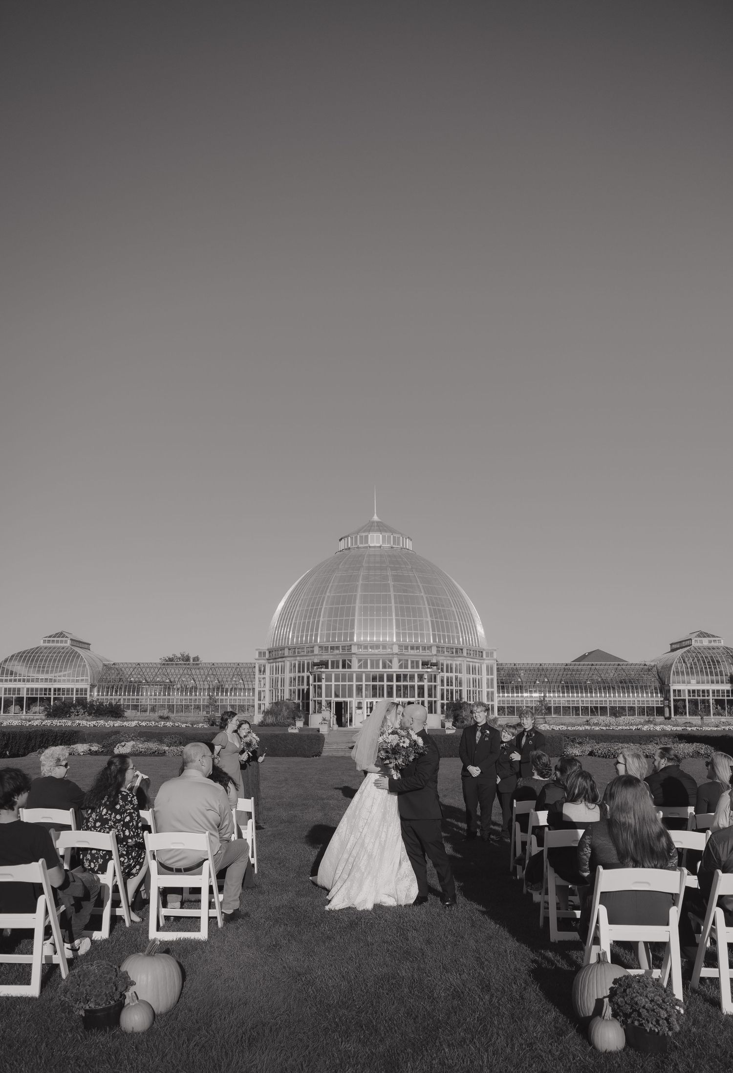 Black and white outdoor wedding ceremony on a lawn with guests seated, facing a grand glass domed conservatory building.