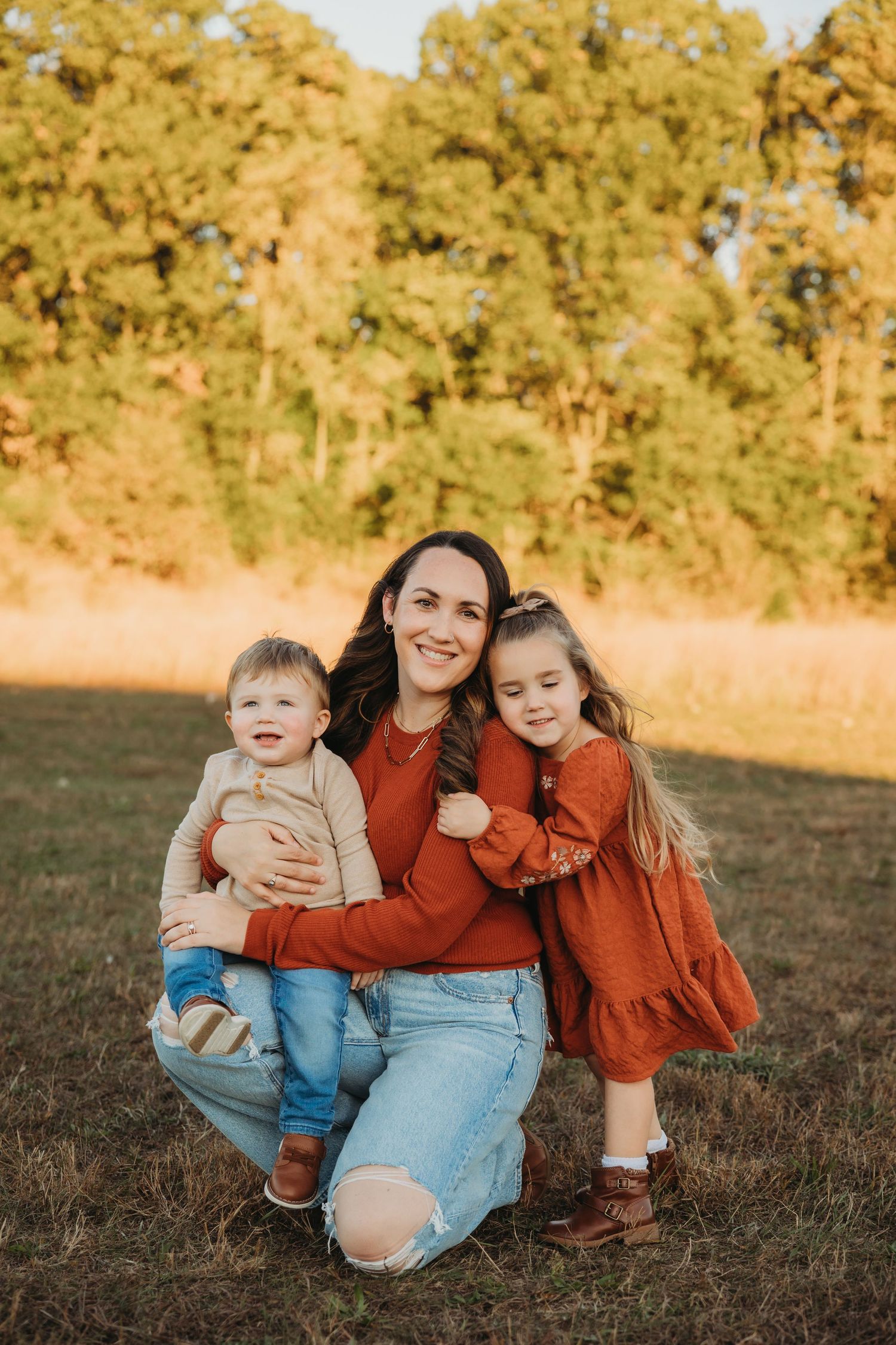 Mother in casual denim kneeling with two children in warm autumn tones against wooded backdrop.