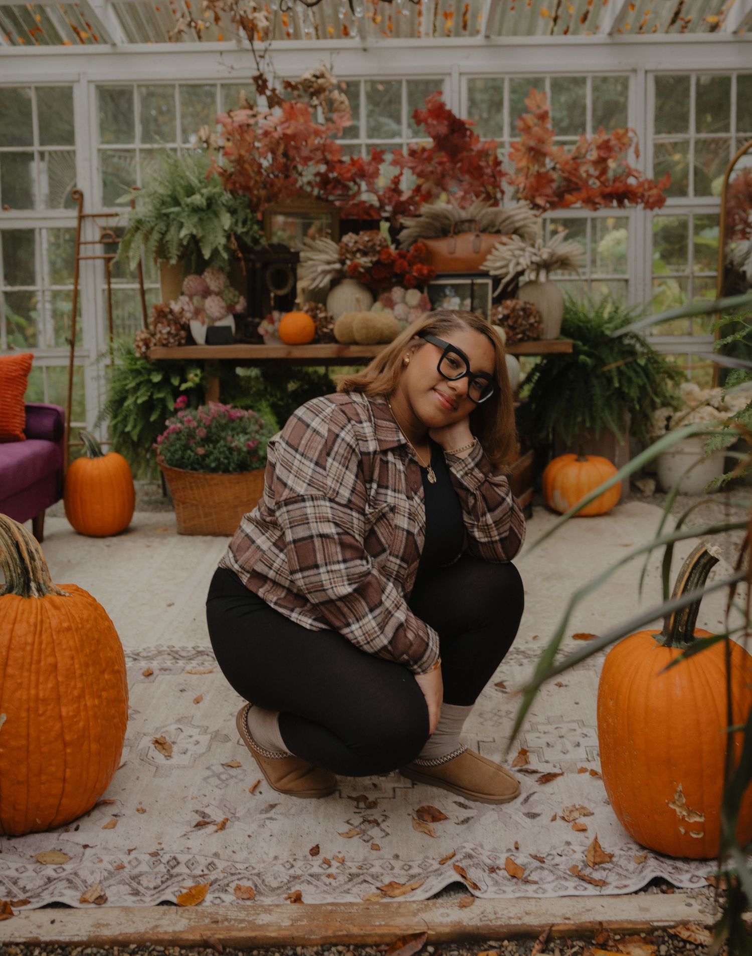 Woman in plaid shirt and glasses crouching among pumpkins in a fall-decorated greenhouse setting.