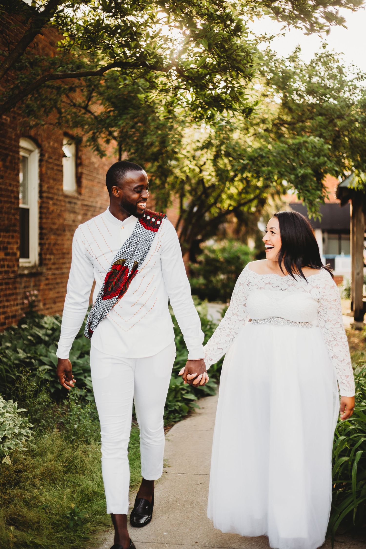 A couple in matching white attire walks hand in hand along a tree-lined brick pathway on a sunny afternoon.