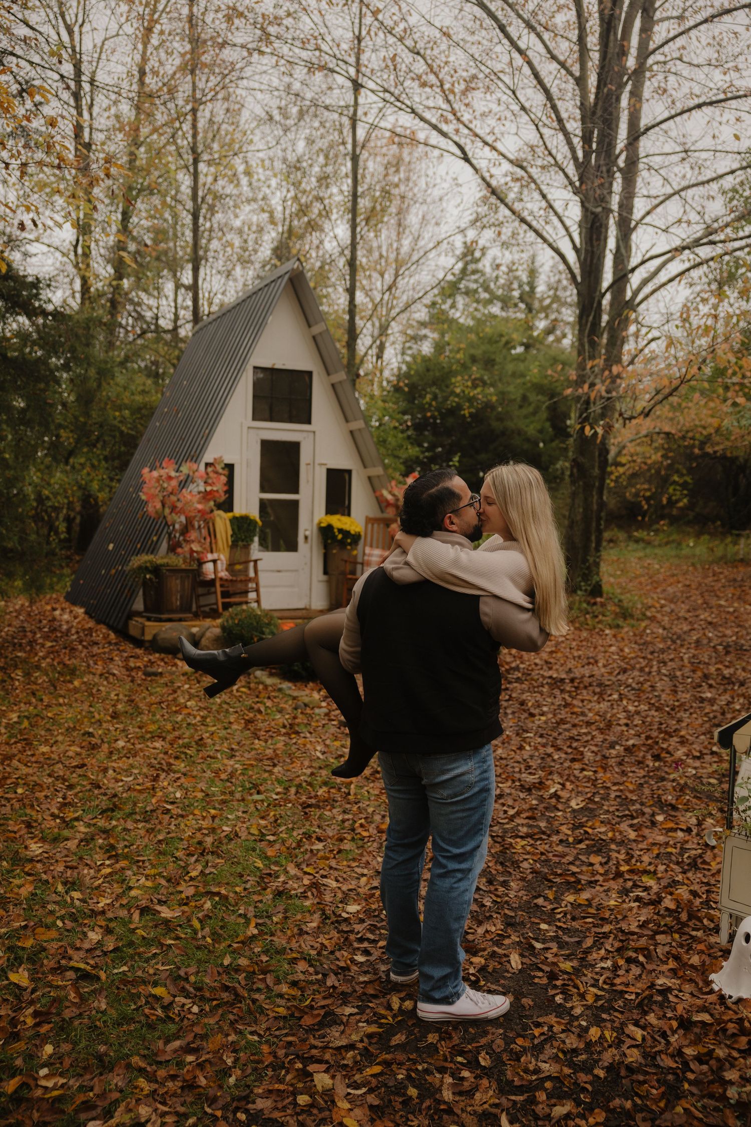 A sequence of small wooden sheds or playhouses stand in a wooded area covered with fallen autumn leaves.