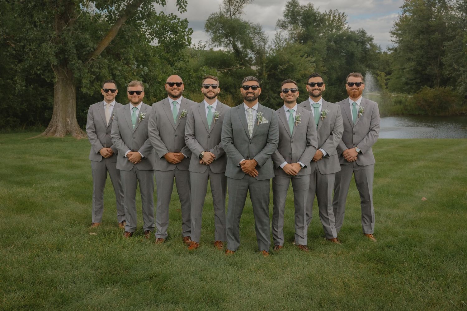 Wedding party in grey suits posing together on grass with trees in background.