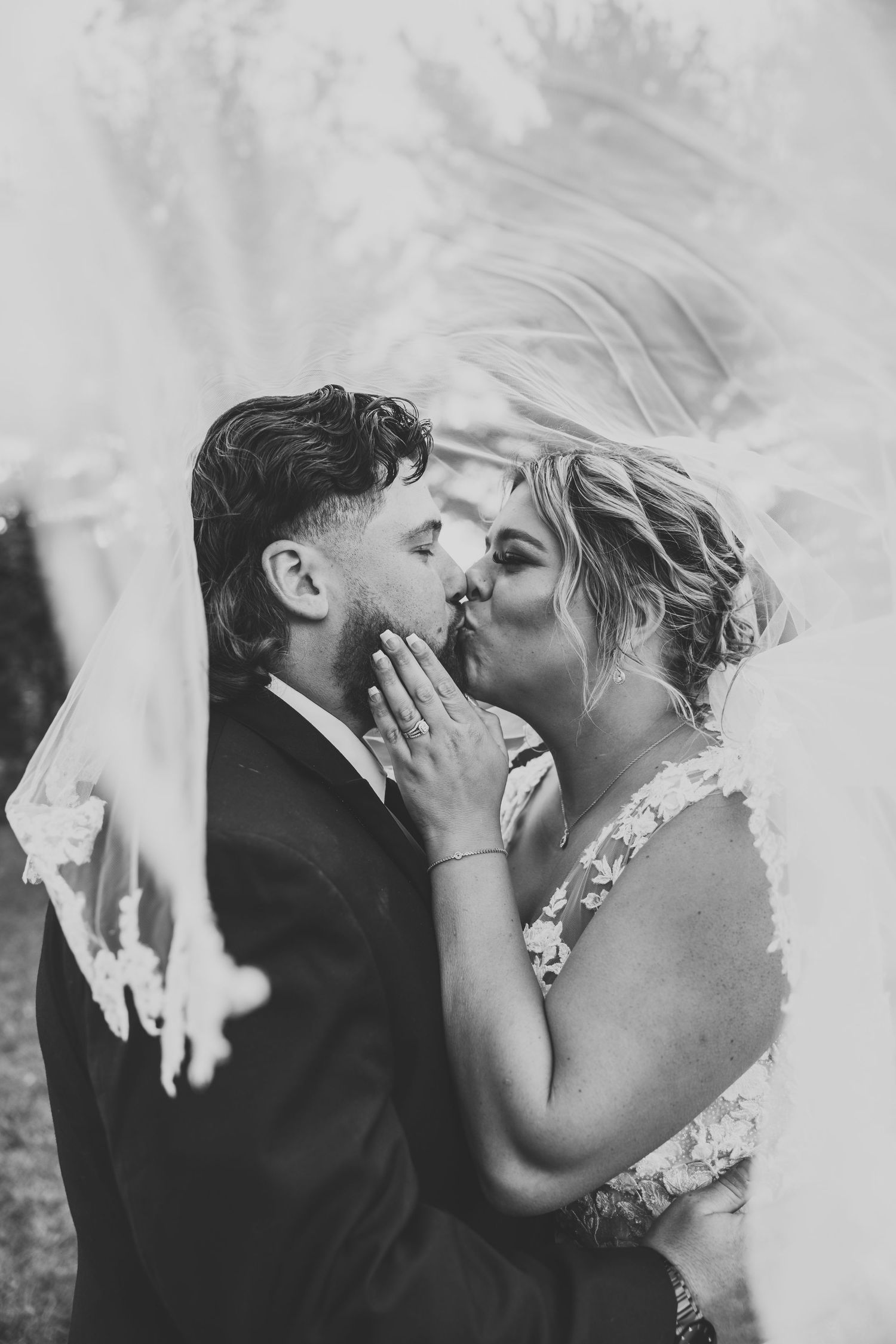Artistic black and white wedding photo showing newlyweds sharing an intimate kiss under a flowing veil.