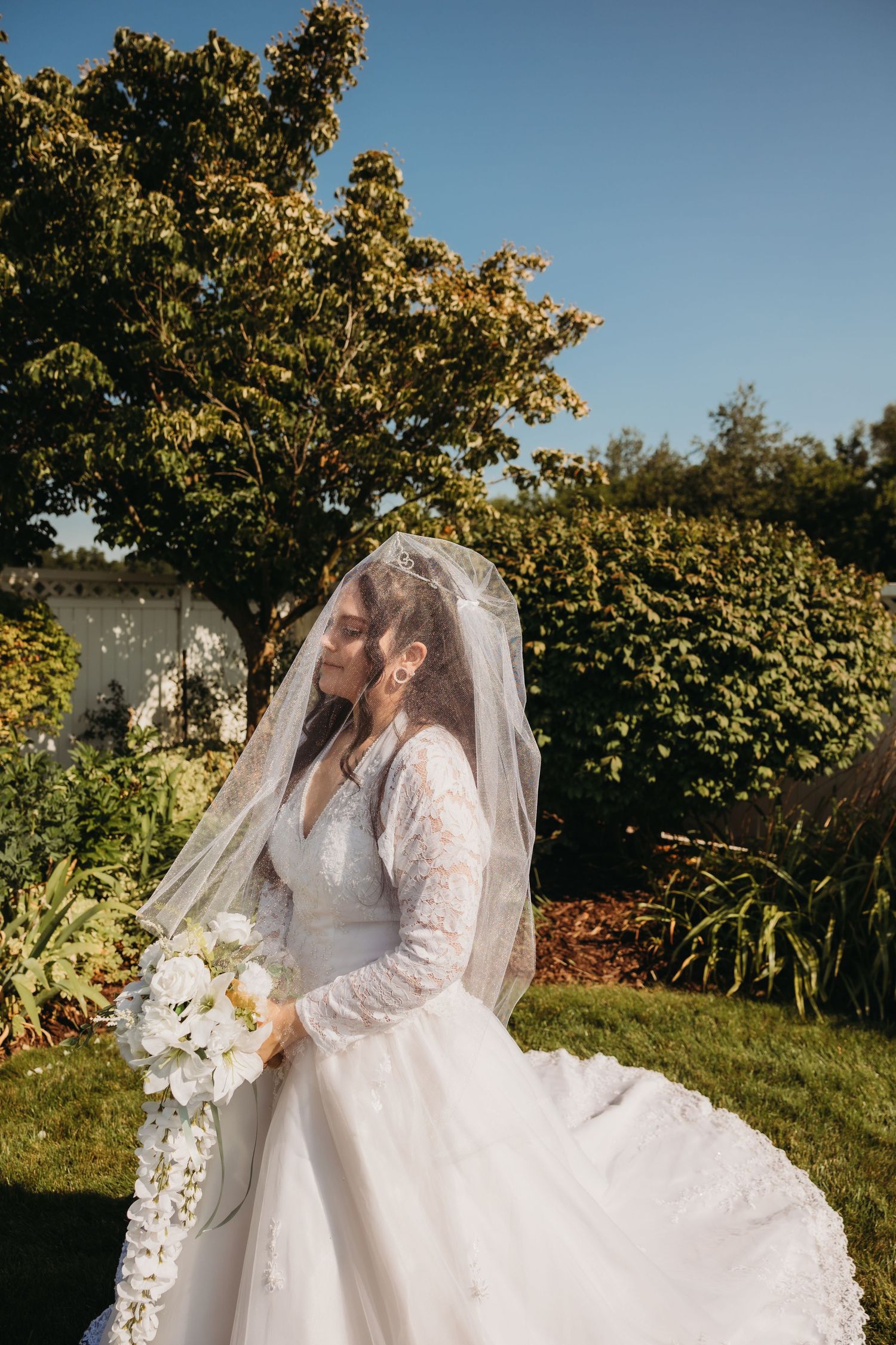 Elegant bride in lace gown and veil holds cascading white bouquet in garden setting.