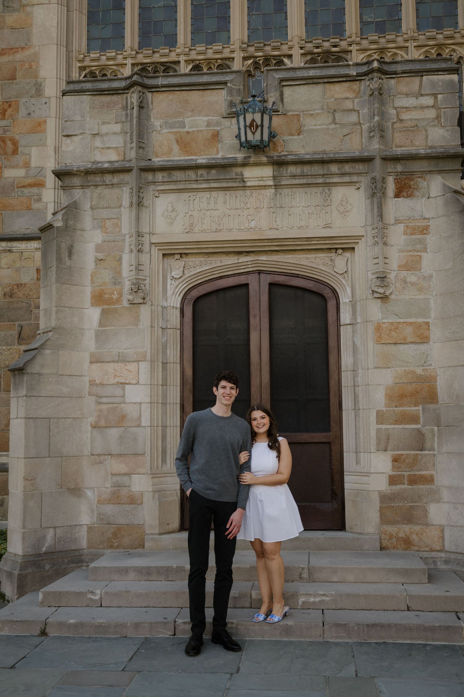 A young couple poses together on stone steps in front of an ornate gothic building with arched wooden doors.