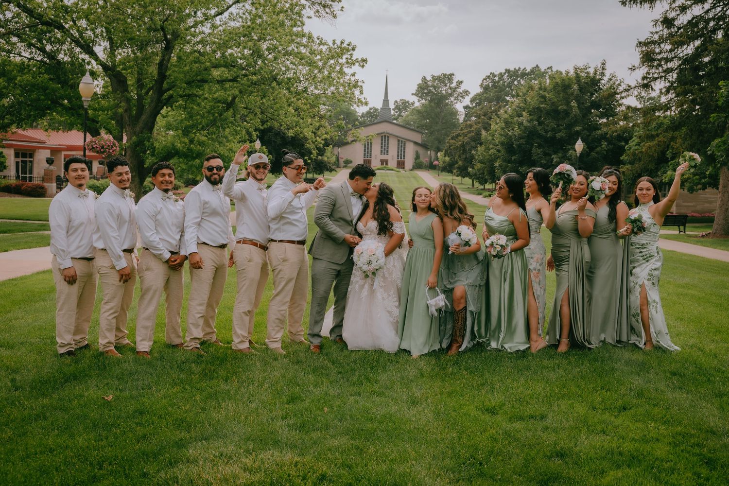 Wedding group photo with bridal party in sage green dresses and groomsmen in khaki suits standing on grass.
