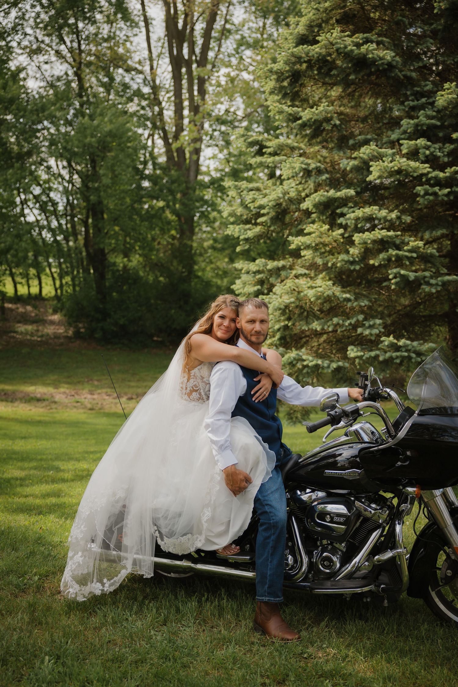 Bride in white gown embraces groom seated on black Harley-Davidson motorcycle on lush green lawn with trees.