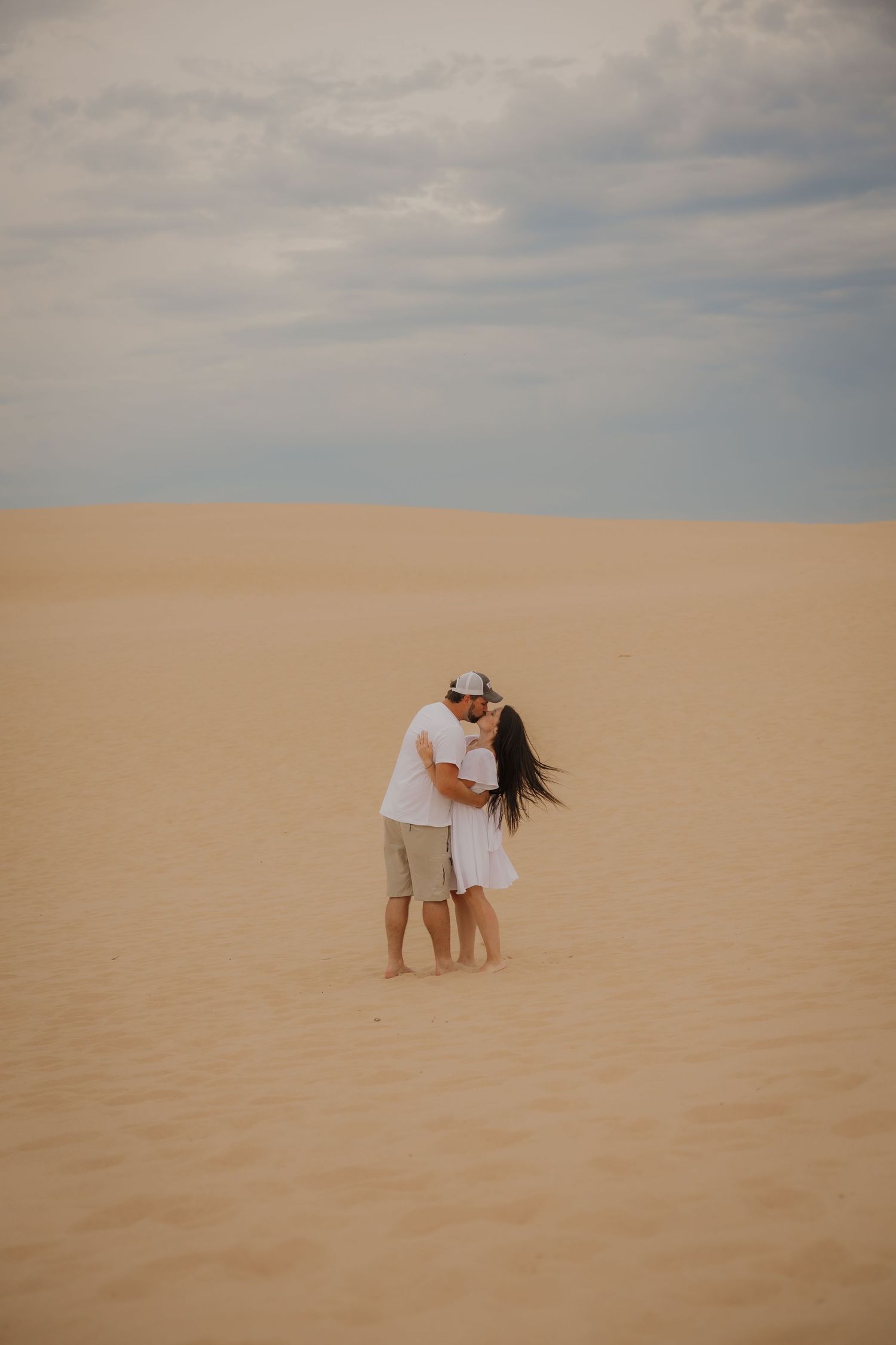 Couple embracing and kissing on vast sandy desert dunes under cloudy sky, wearing white outfits.