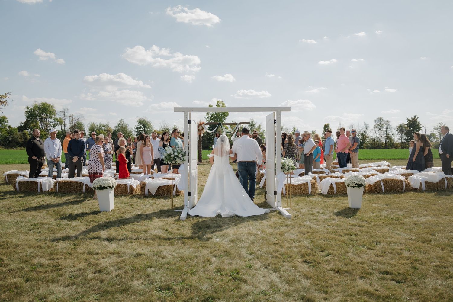 Outdoor wedding ceremony on green lawn with bride and groom at white arch, guests seated on hay bales under blue sky.