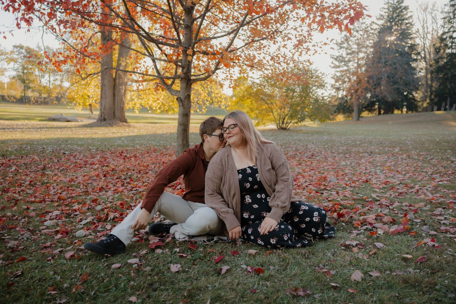 Couple sitting on fall leaves under colorful autumn trees in a park, sharing a tender romantic moment.