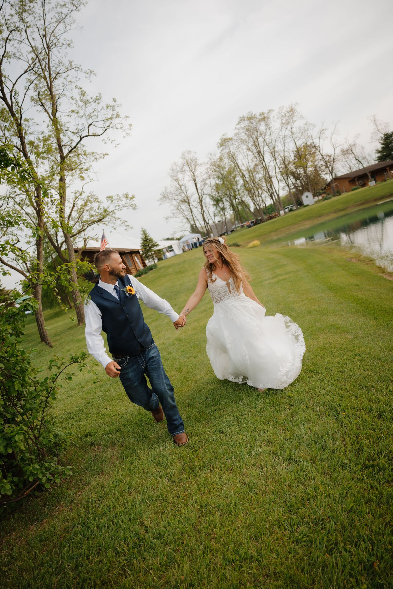 Bride and groom holding hands running on green grass near a pond on their wedding day.