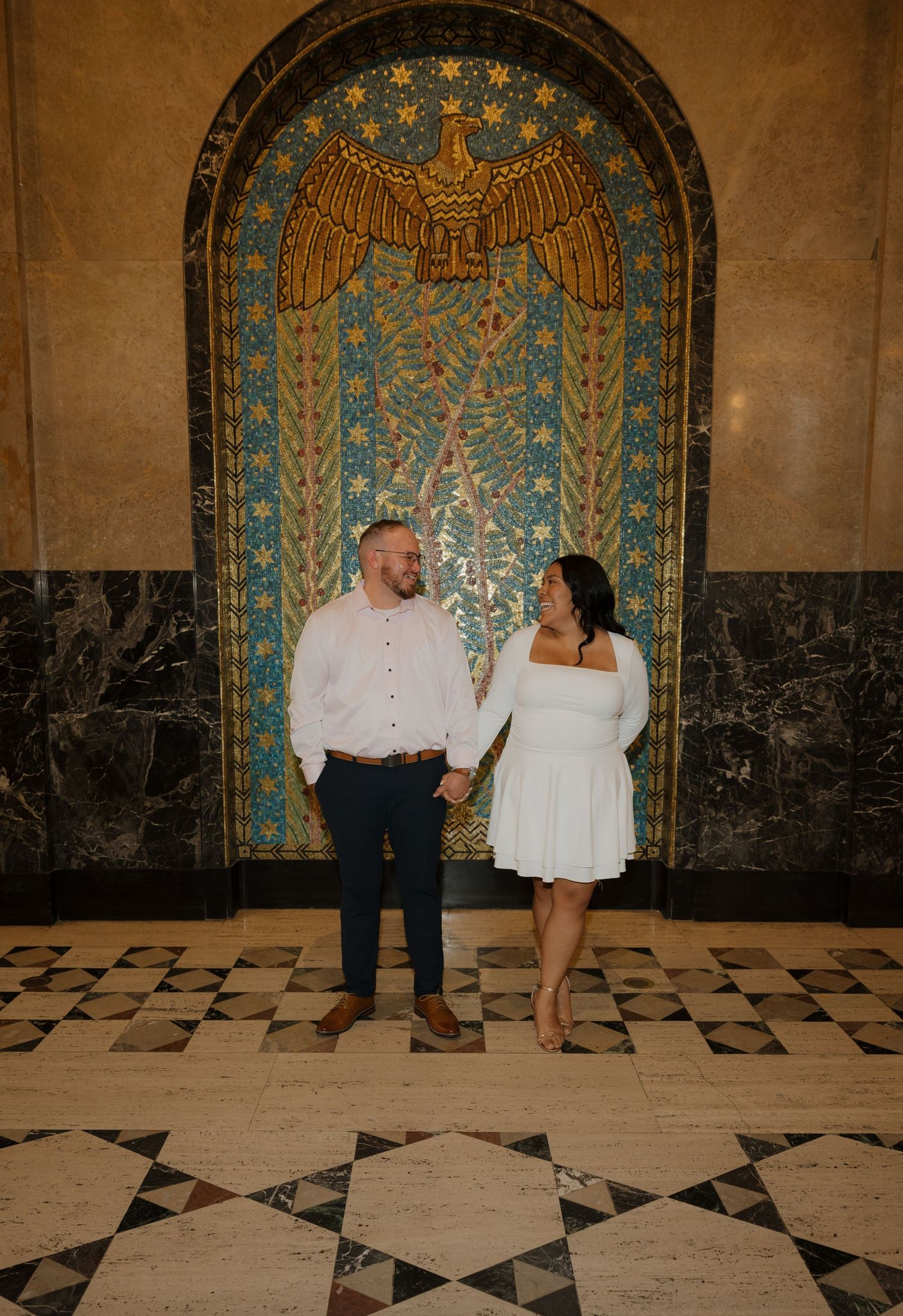 Couple holding hands smiling at each other in front of ornate mosaic mural with eagle design on marble wall.