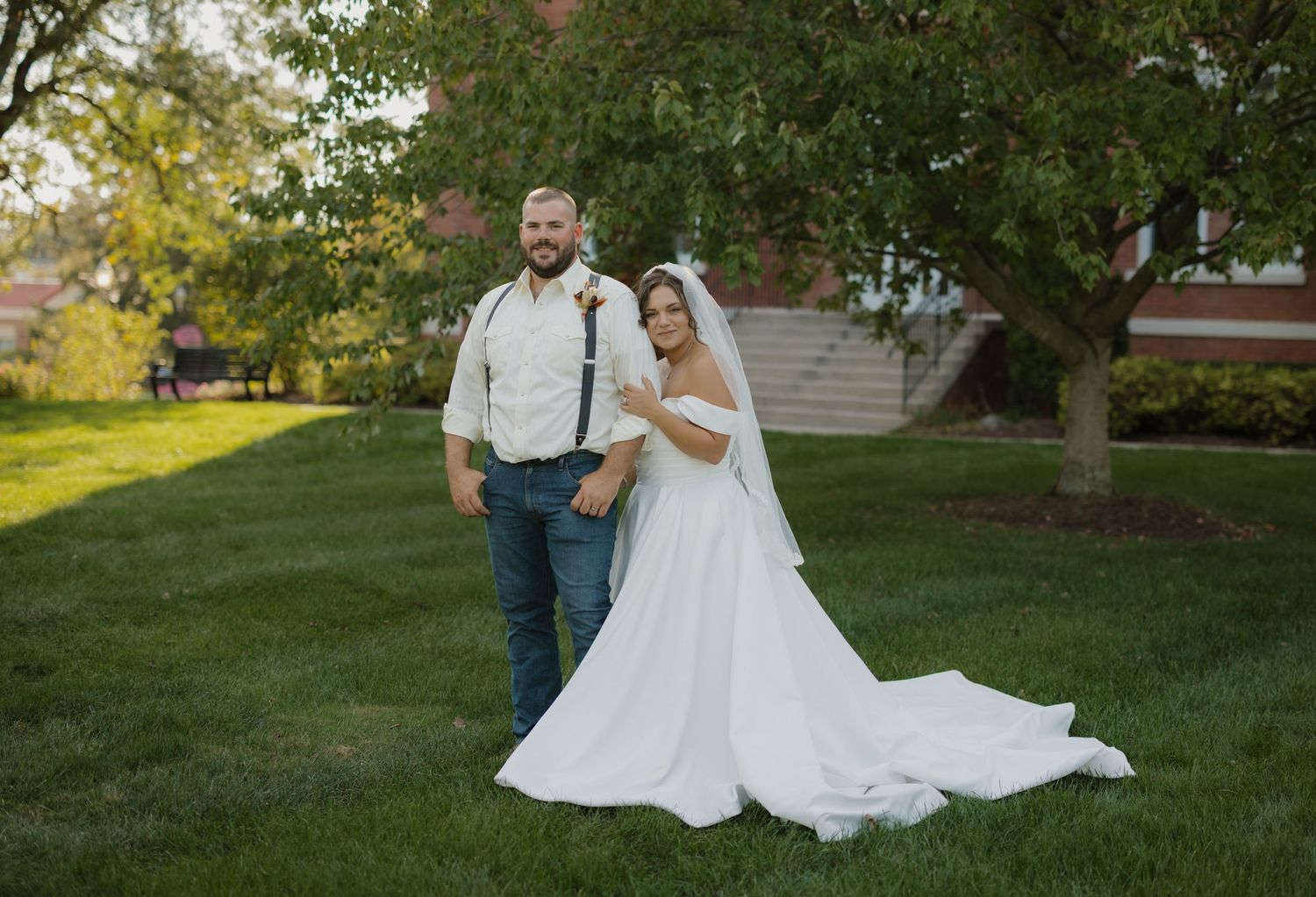 Bride in white gown and groom in jeans and suspenders pose together on green lawn under tree.