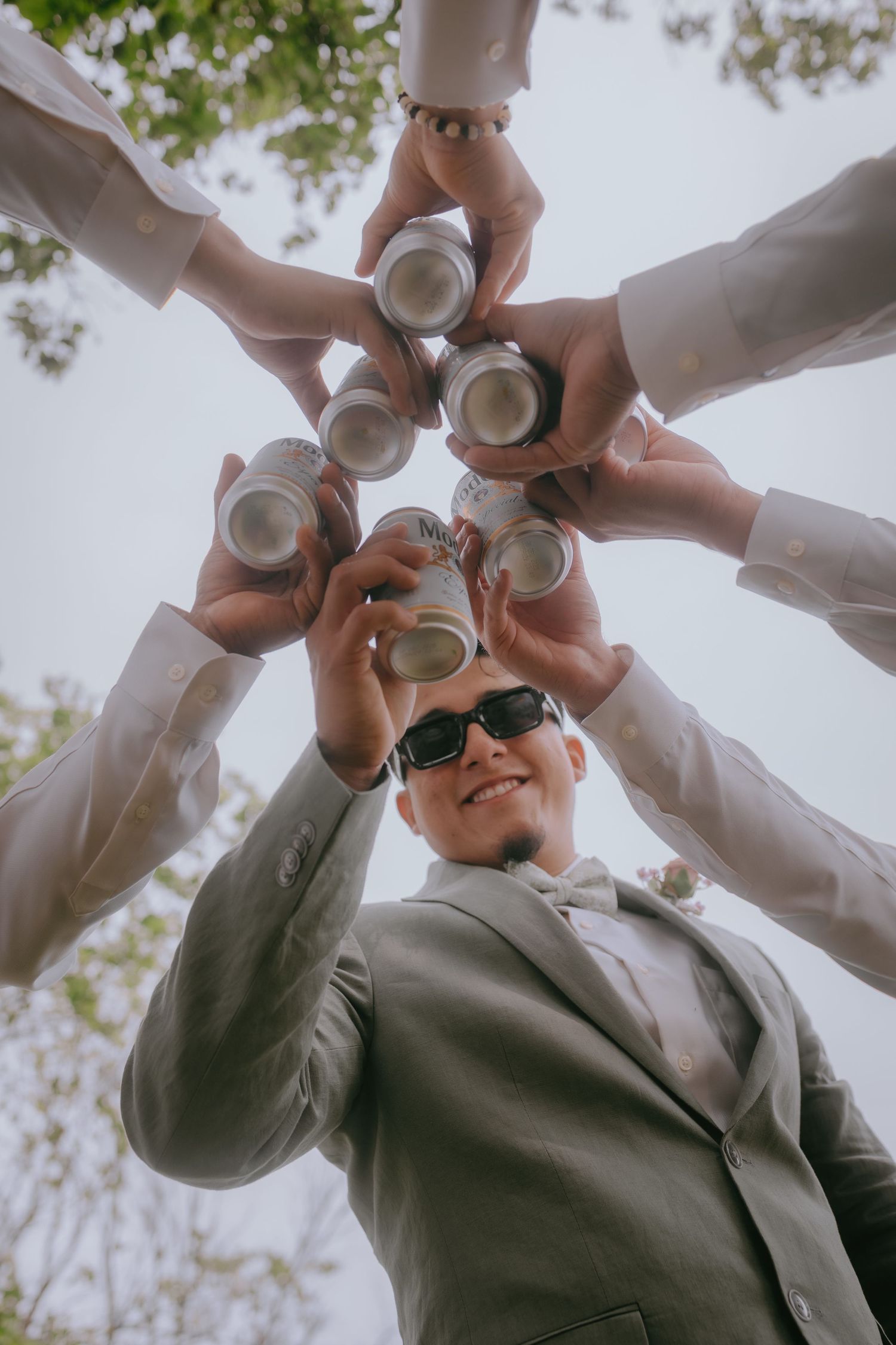 A group toasts with aluminum cans in a circular formation photographed from below against a bright sky.