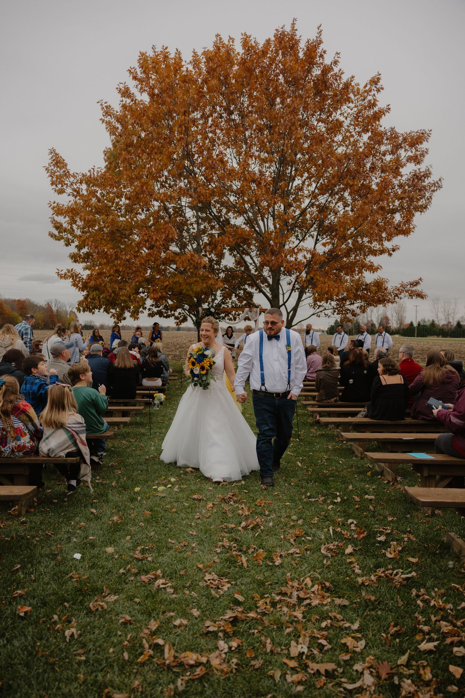 Bride and groom walk down outdoor aisle lined with wooden benches beneath a vibrant autumn tree with orange foliage.