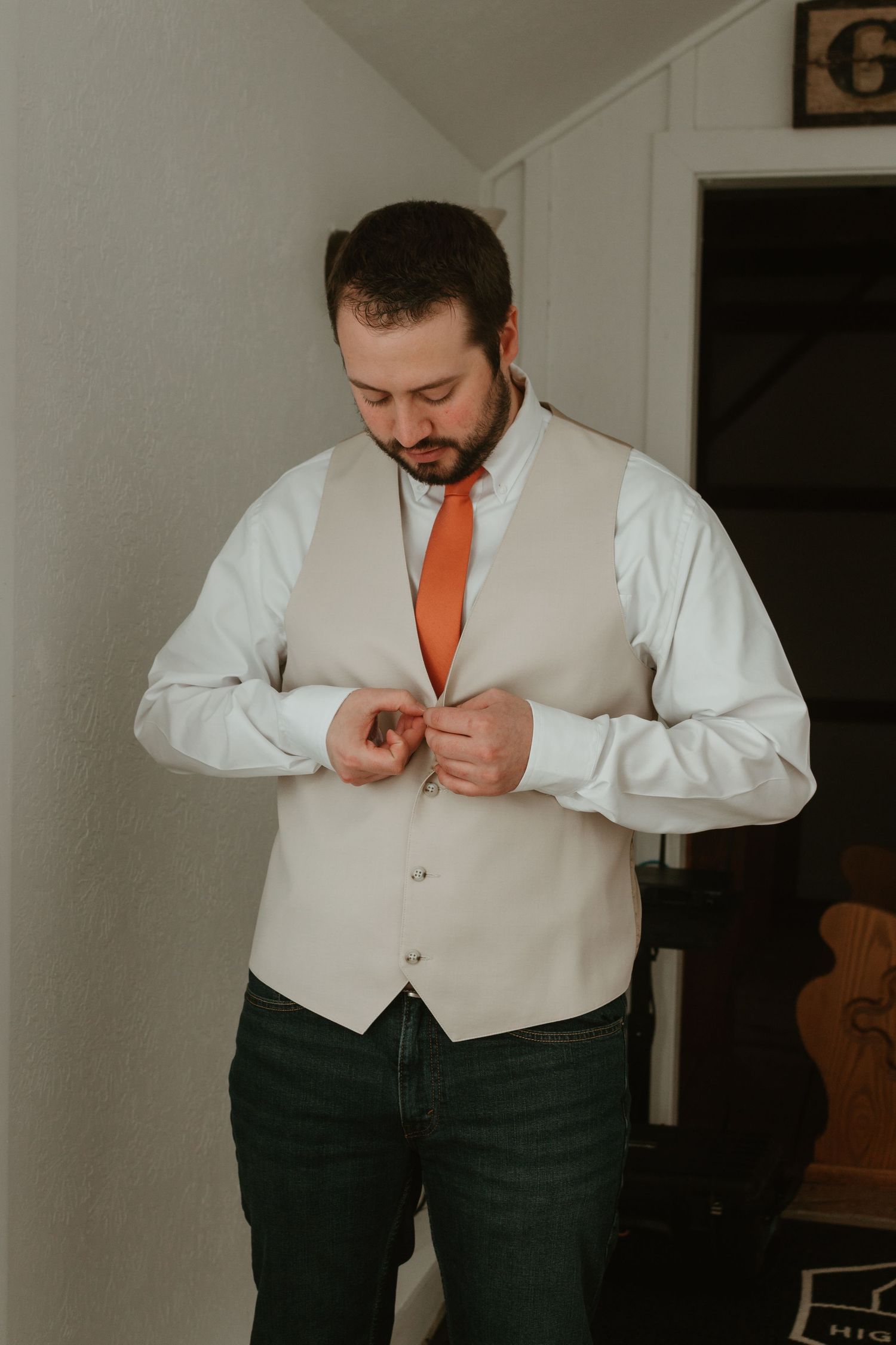 A groom in a beige vest and orange tie adjusts his buttons while getting ready.