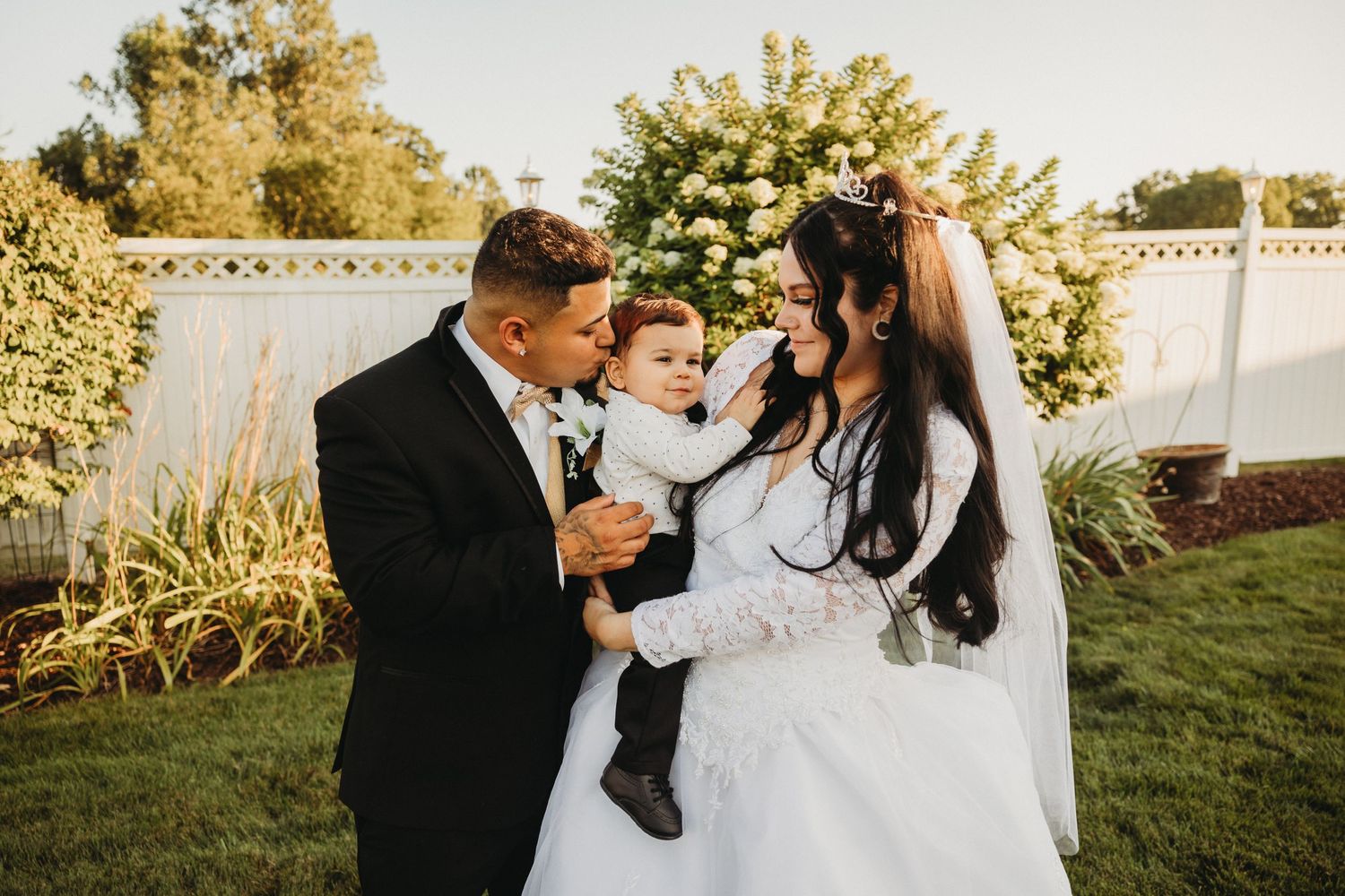 Wedding couple outdoors holding a small child against a white fence and garden backdrop.