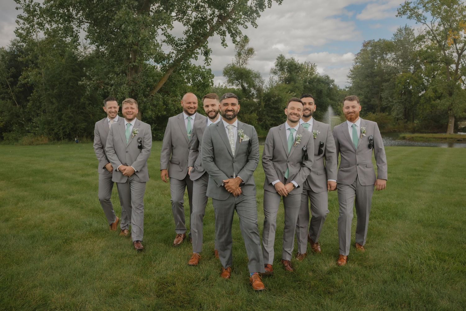 Groomsmen in gray suits with brown shoes lined up for formal wedding photos outdoors.