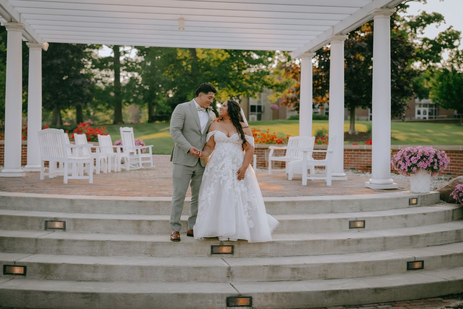 A couple celebrates their wedding day on stone steps under a white gazebo with chairs set up in the background.