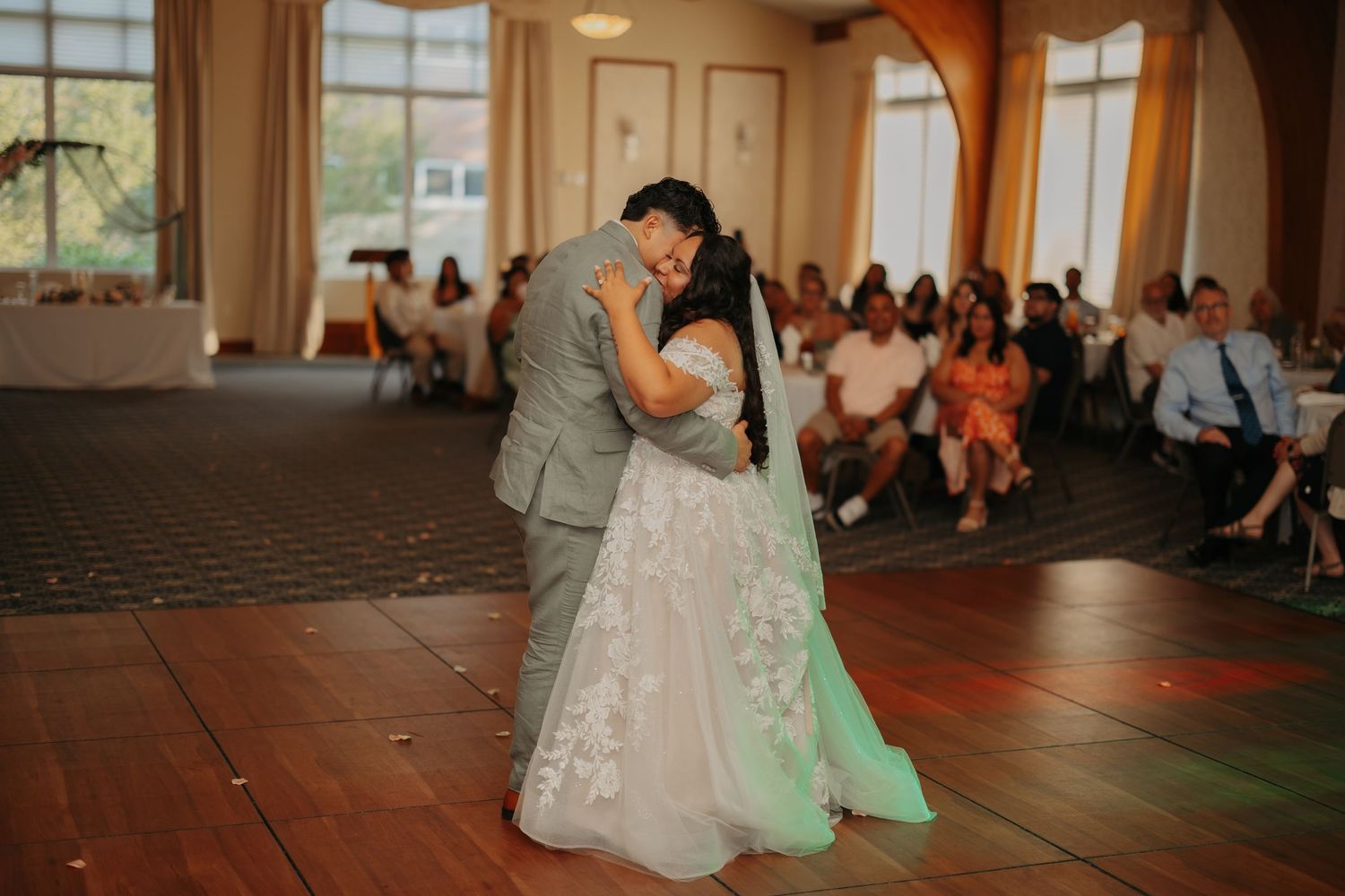A couple shares their first dance as newlyweds on a wooden floor while wedding guests look on from their seats.
