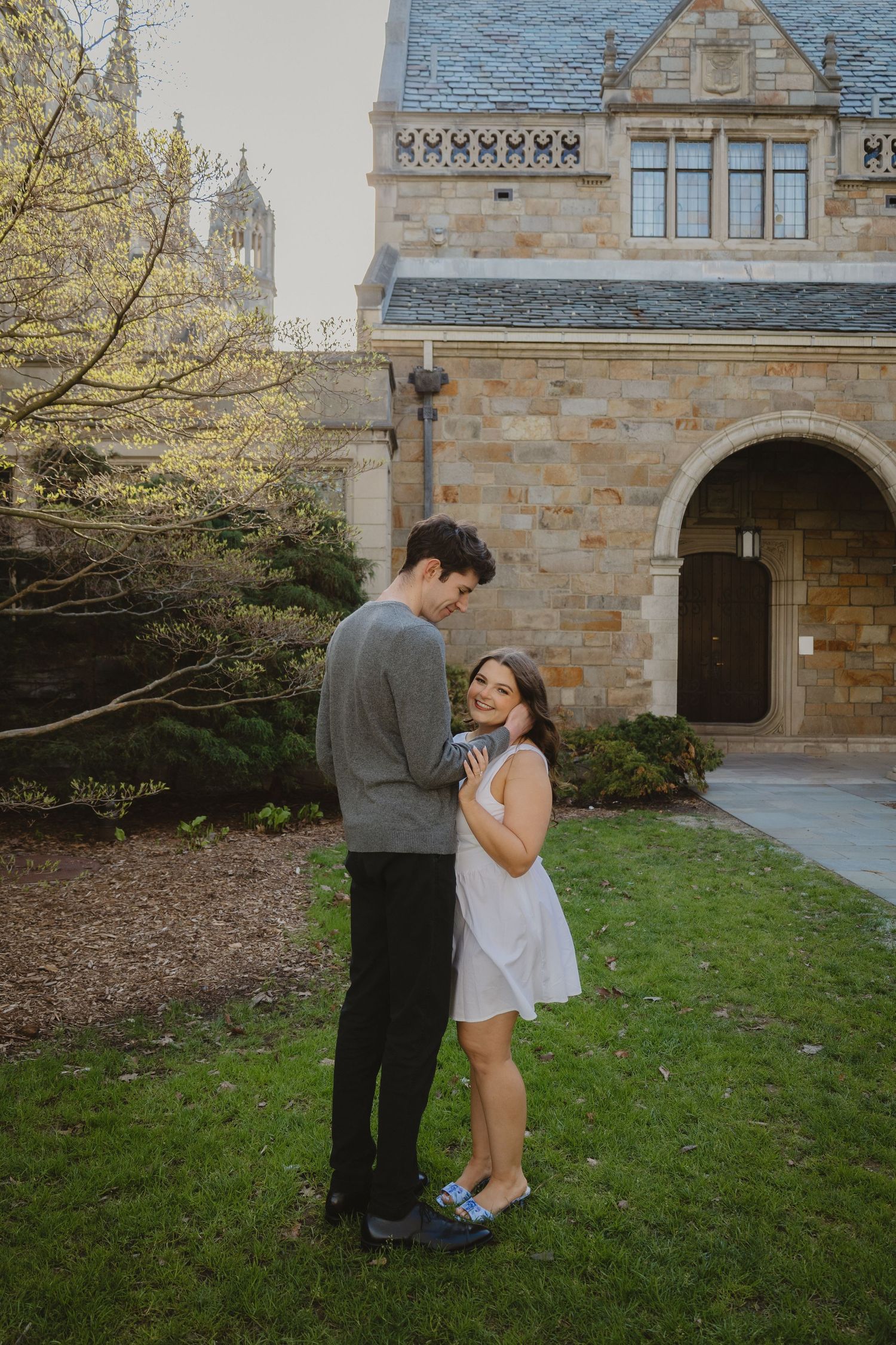 Couple embracing on grass lawn in front of a stone building, woman in white dress smiling up at man in gray sweater.