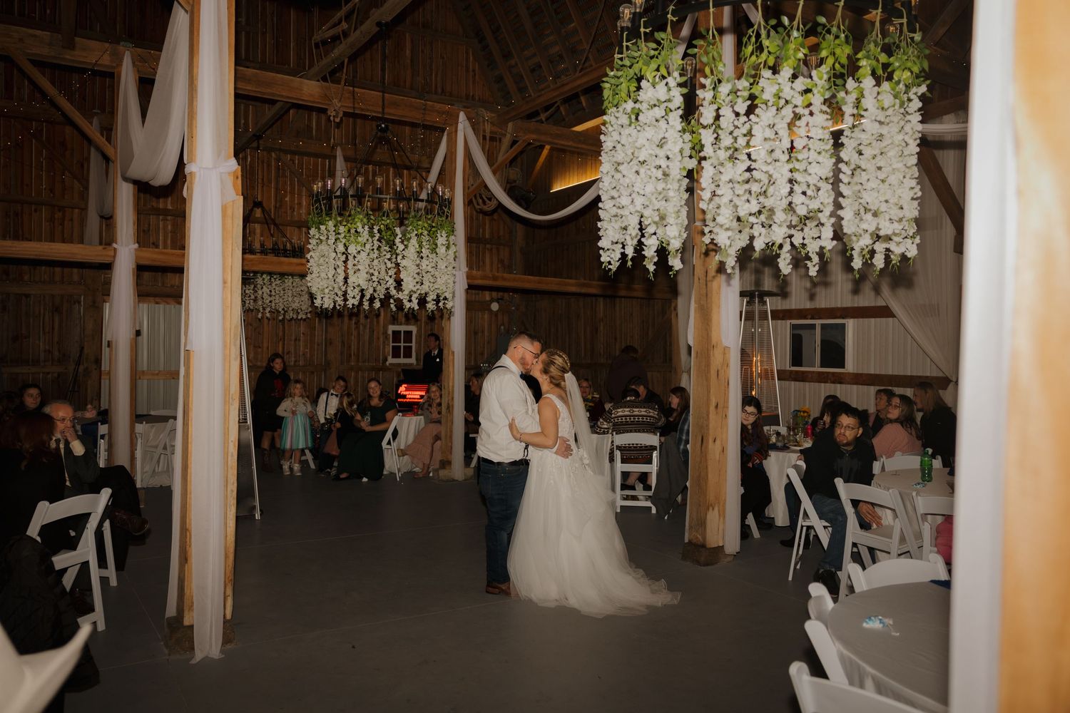 Bride and groom share first dance in a rustic barn venue adorned with hanging floral chandeliers and white draping.